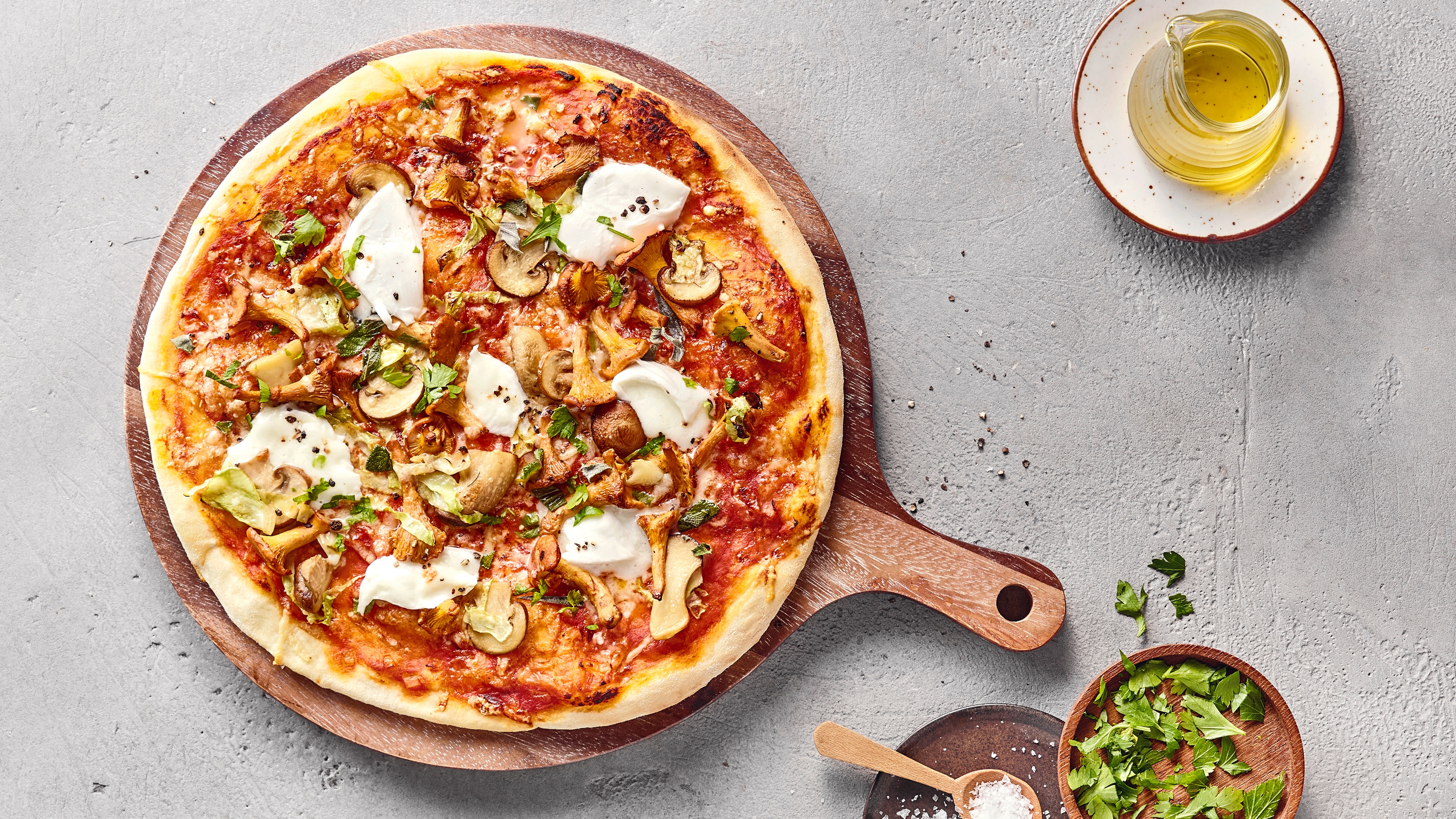 Pizza mit Tomatensoße, Käse, Pilzen und Mozzarella auf einem Holzbrett, daneben Olivenöl und frische Kräuter