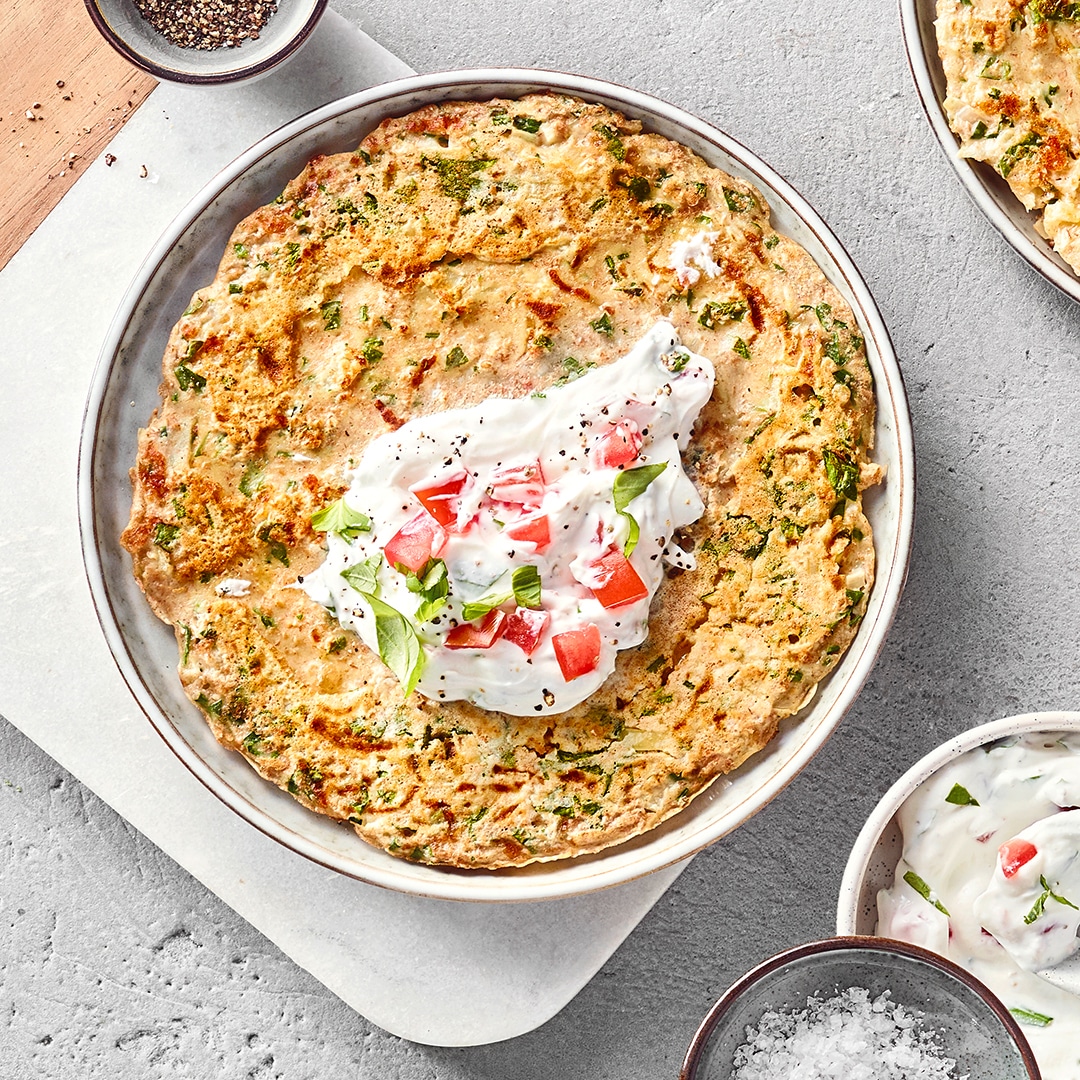 Ein Teller mit gelbgoldenen Pfannkuchen mit Quark-Joghurt-Dip, Tomaten und frischen Kräutern
