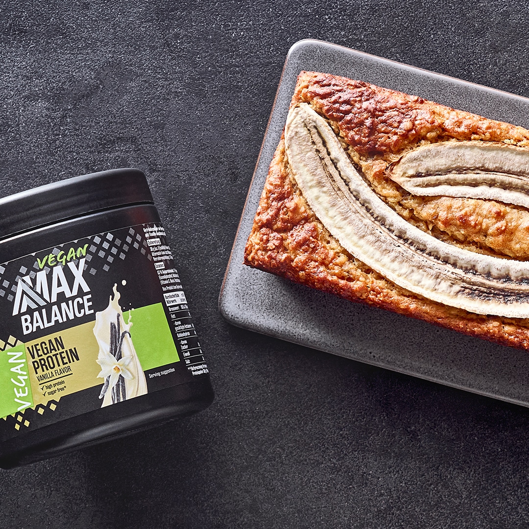 Kastenförmiger Kuchen mit eingebackenen Bananenhälften auf Brett, daneben eine Dose MaxBalance Veganes Proteinpulver Vanille