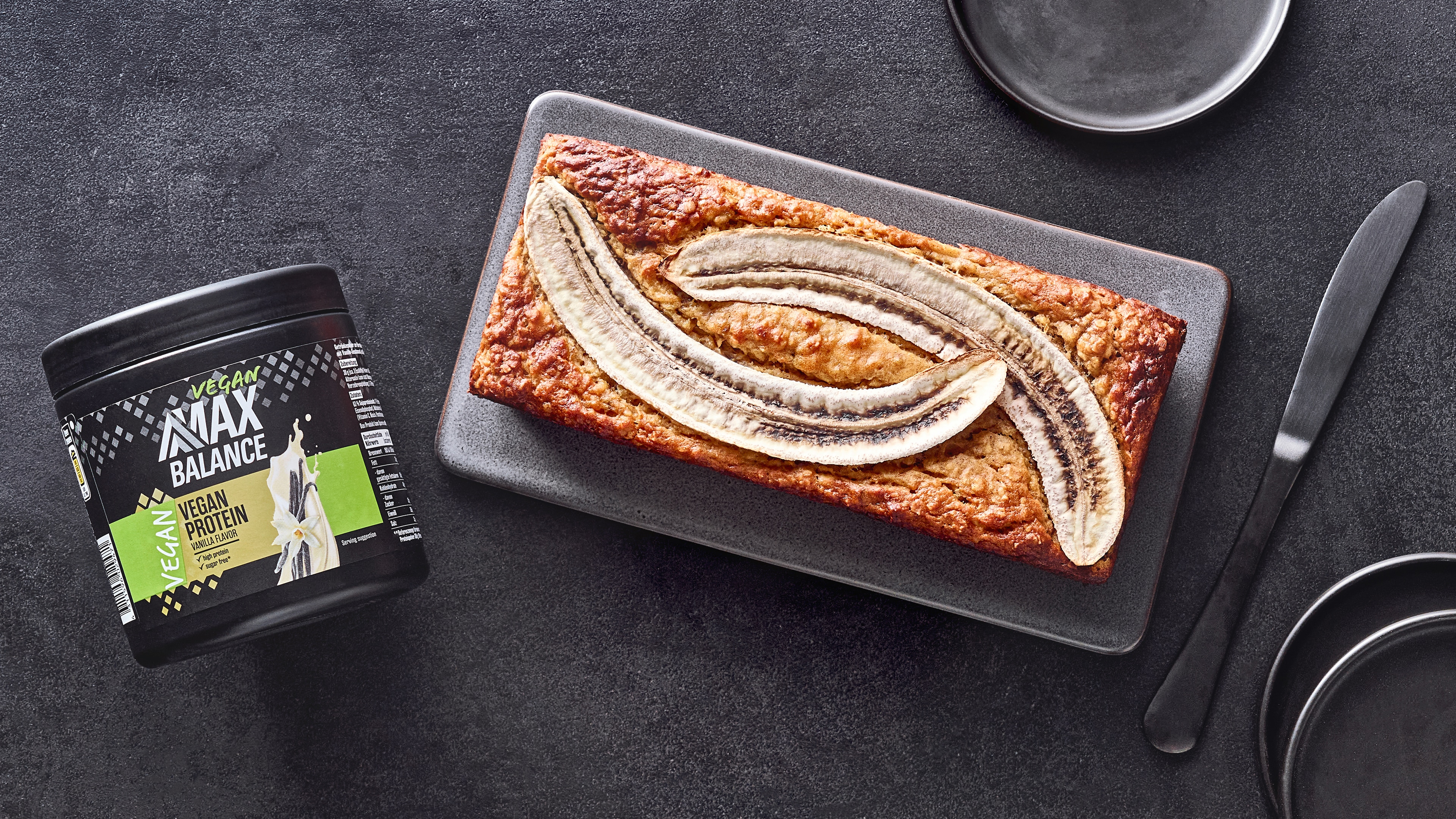 Kastenförmiger Kuchen mit eingebackenen Bananenhälften auf Brett, daneben eine Dose MaxBalance Veganes Proteinpulver Vanille
