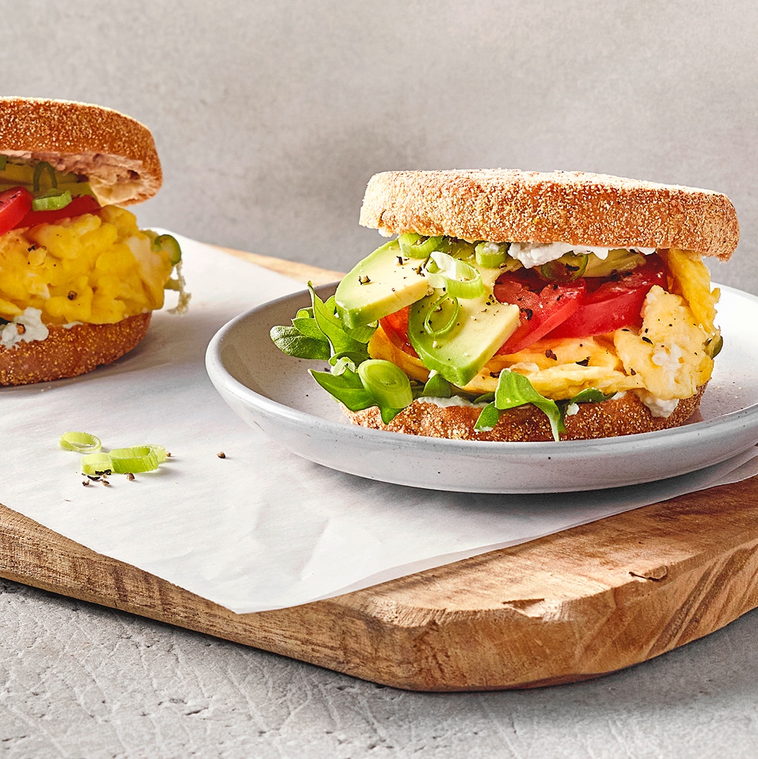 Belegte Frühstücksbrötchen mit Rührei, Avocado, Tomate und Salat auf Tellern und einem Holzbrett angerichtet.