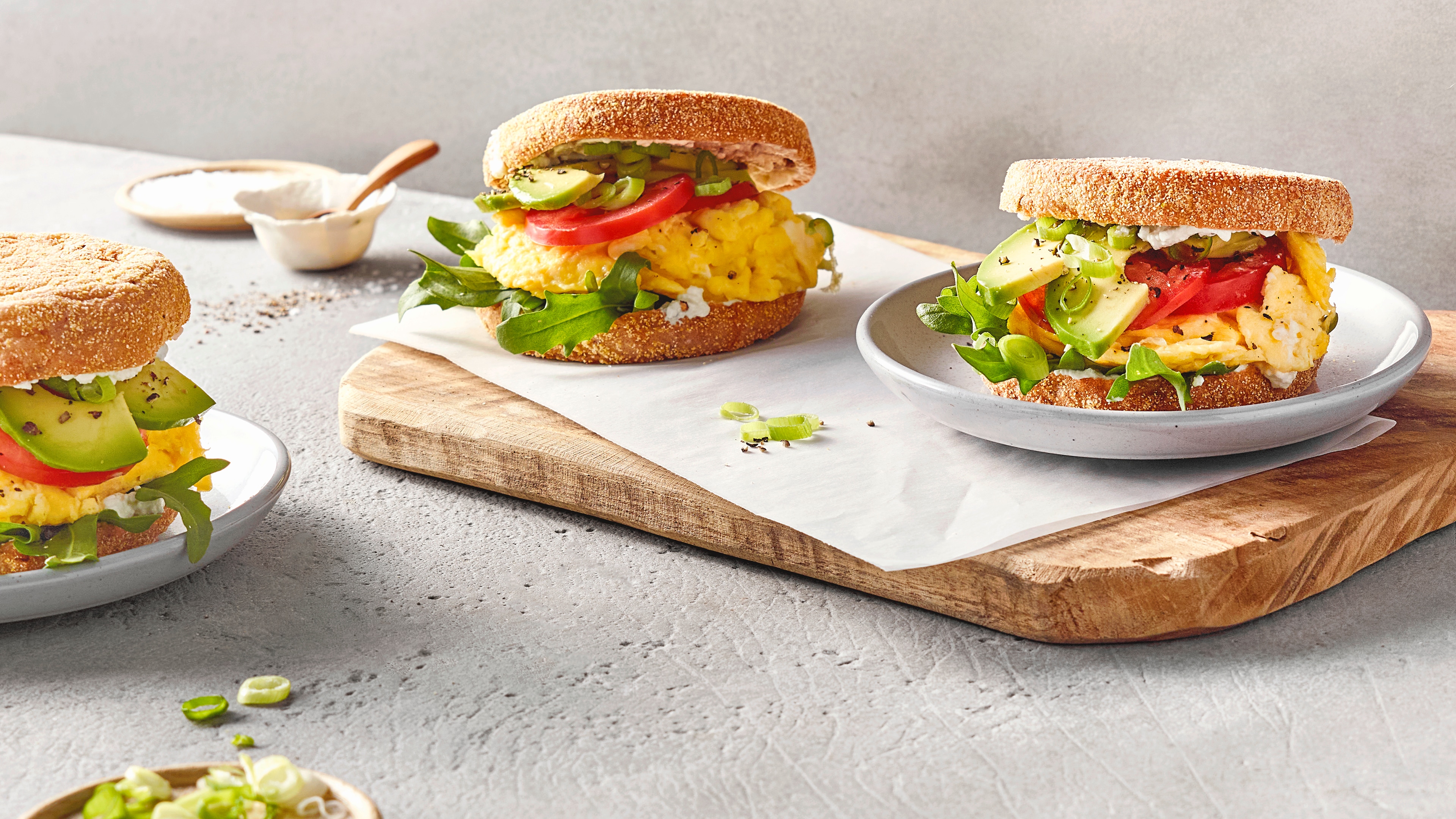 Drei belegte Frühstücksbrötchen mit Rührei, Avocado, Tomate und Salat auf Tellern und einem Holzbrett angerichtet.
