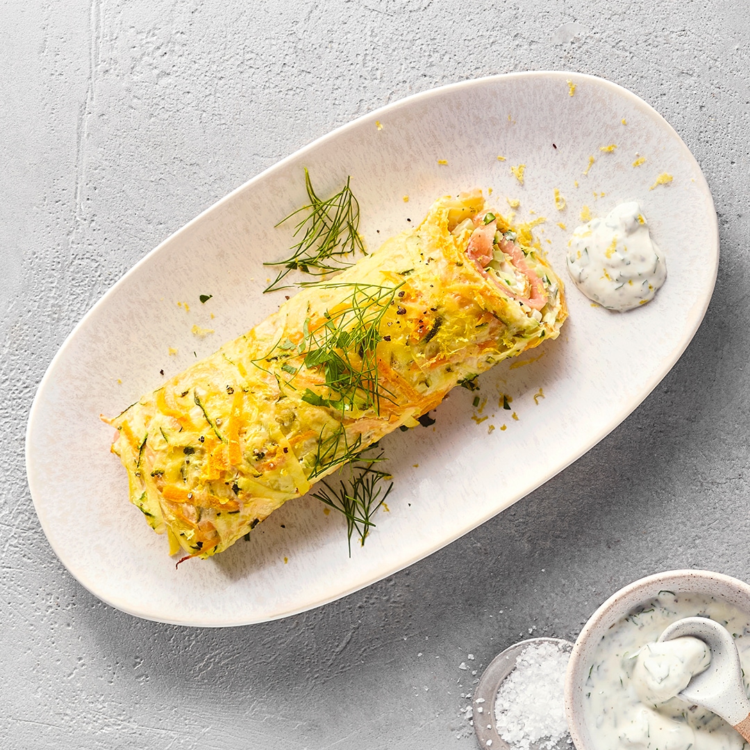 Gemüserolle mit Lachs und Kräuterfrischkäse, garniert mit Dill, daneben eine Schale Kräuterfrischkäse.