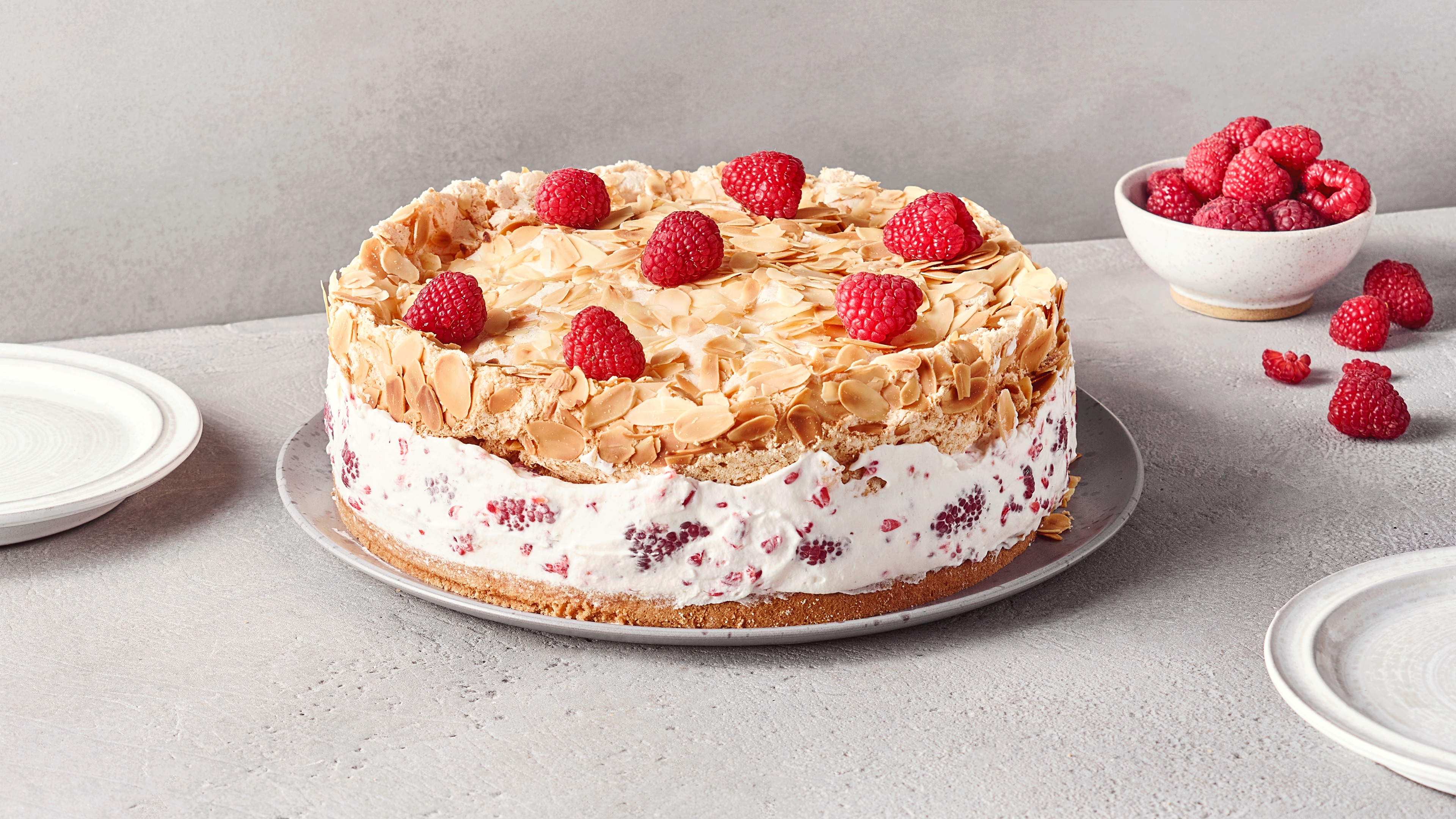 Ein Teller mit einem runden Kuchen, weißer Creme mit Himbeeren, einer Schicht aus Mandelblättchen & roten Himbeeren obenauf.