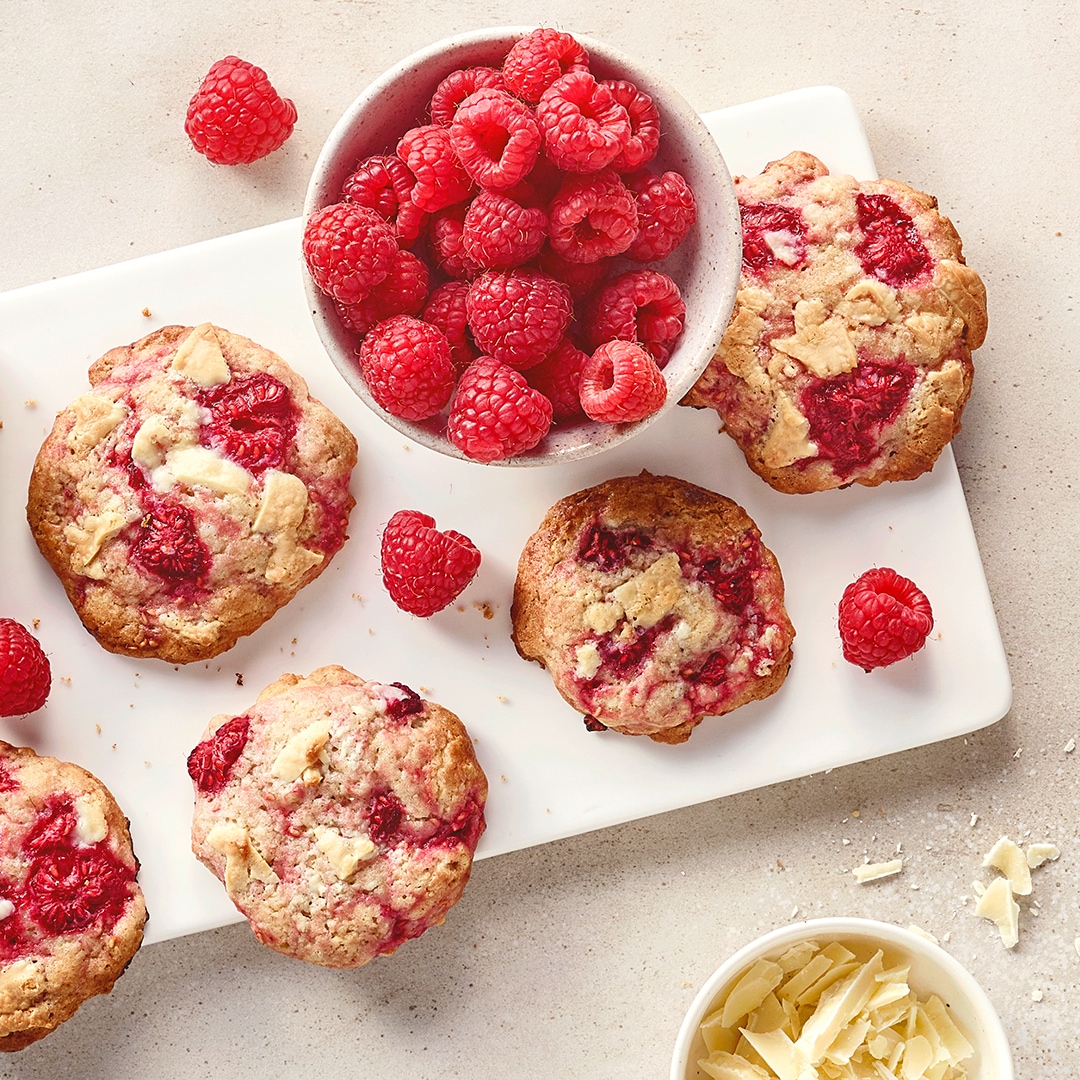 Ein Brett mit Keksen, gefüllt mit Himbeeren und weißen Schokoladenstücken, daneben eine Schale voller frischer Himbeeren.