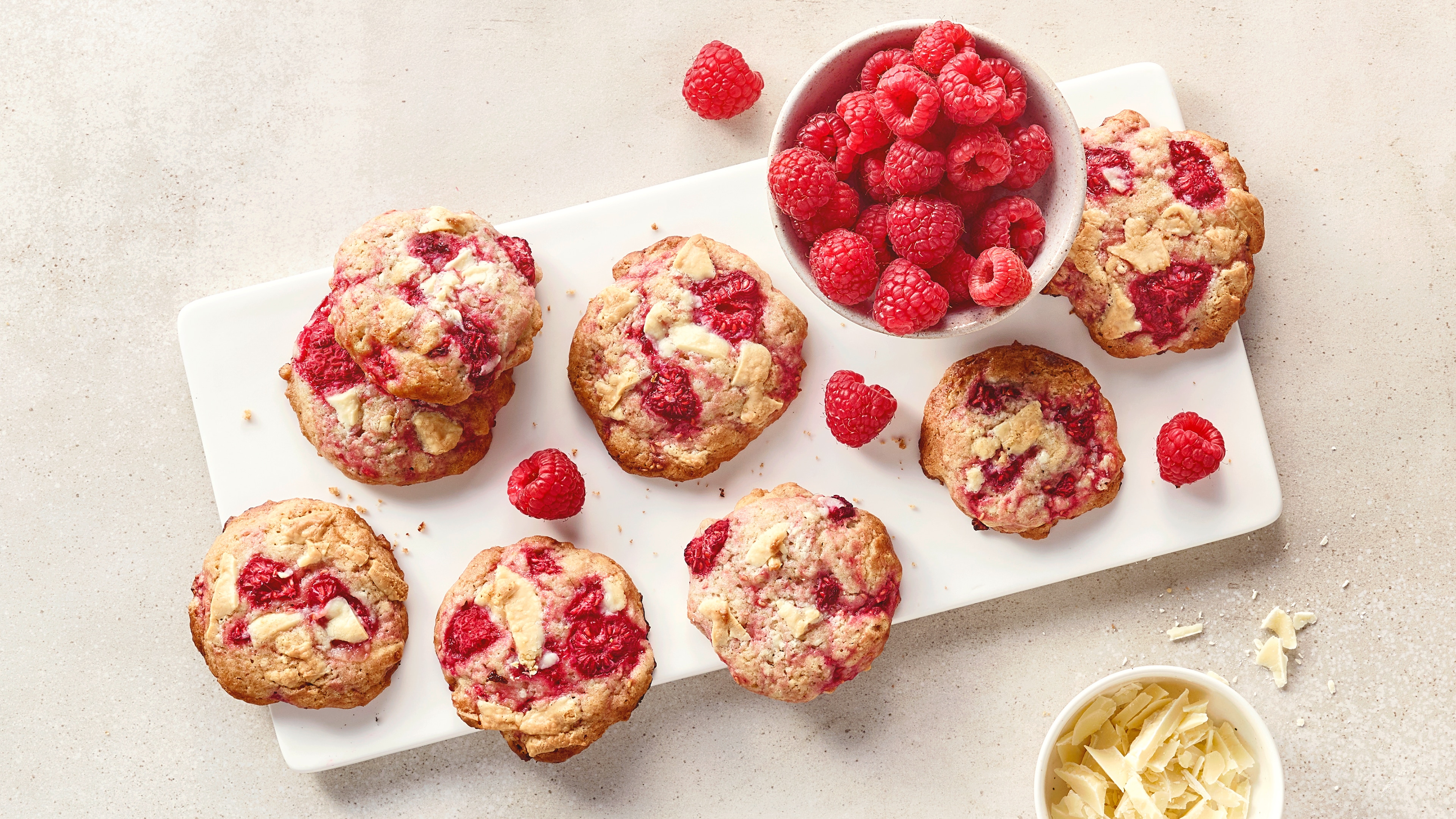 Ein Brett mit Keksen, gefüllt mit Himbeeren und weißen Schokoladenstücken, daneben eine Schale voller frischer Himbeeren.