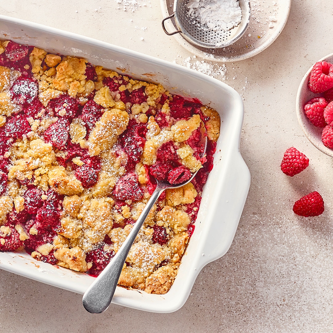 Eine Keramikauflaufform mit gelbgold gebackenen Teigstreuseln und saftigen Himbeeren, bestreut mit feinem Puderzucker