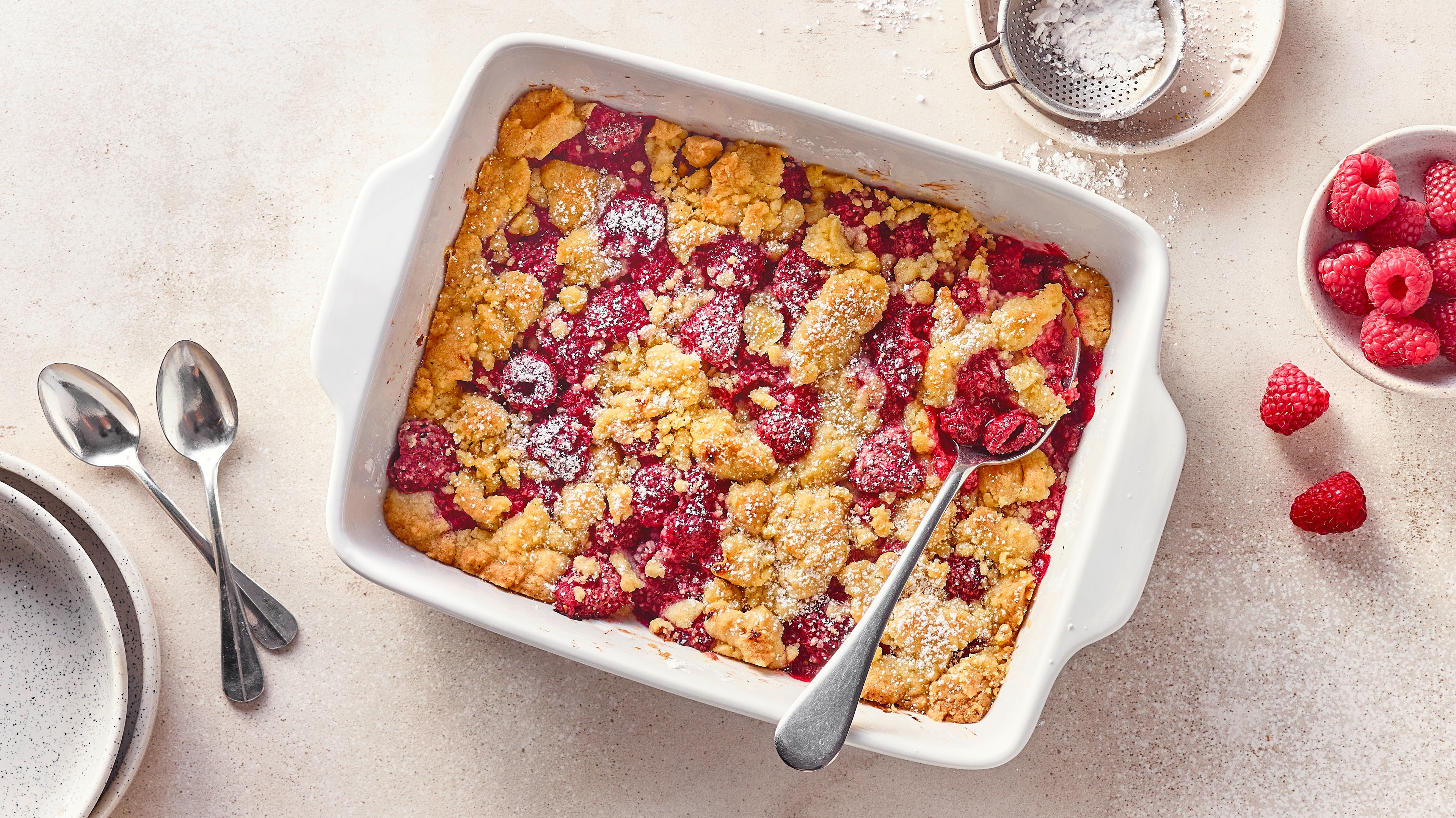 Rezept für Himbeer-Crumble Eine Keramikauflaufform mit gelbgold gebackenen Teigstreuseln und saftigen Himbeeren, bestreut mit feinem Puderzucker