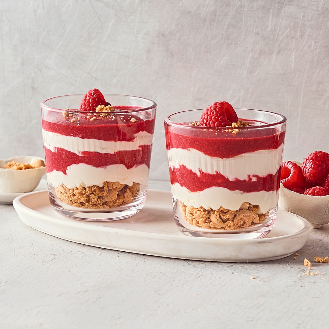 Dessertgläser, befüllt mit Keksstückchen, darüber geschichtet helle und himbeerfarbige Creme, garniert mit frischen Himbeeren