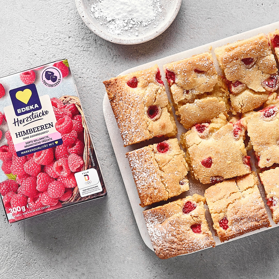 Helle, rechteckige Kuchenstücke mit Himbeeren und Puderzucker auf Brett. Links daneben EDEKA Herzstücke Himbeeren.