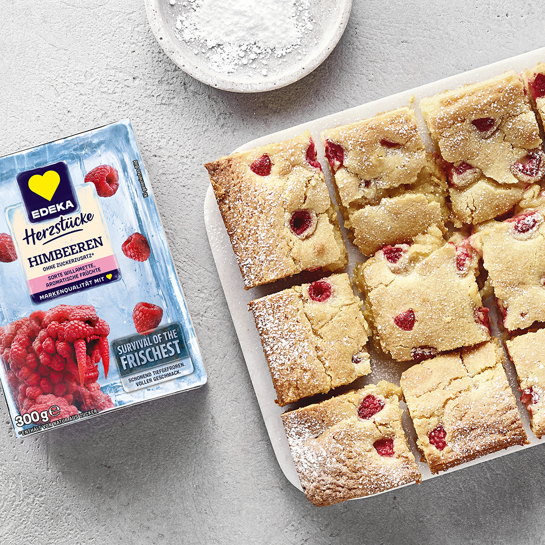 Helle, rechteckige Kuchenstücke mit Himbeeren und Puderzucker auf Brett. Links daneben EDEKA Herzstücke Himbeeren.
