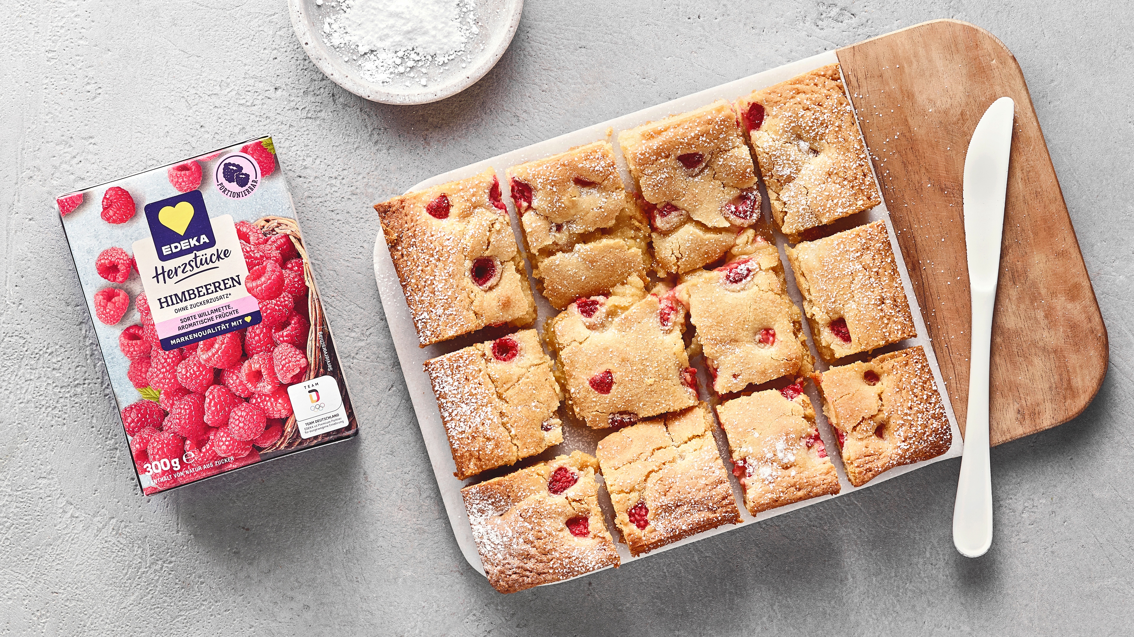 Helle, rechteckige Kuchenstücke mit Himbeeren und Puderzucker auf Brett. Links daneben EDEKA Herzstücke Himbeeren.