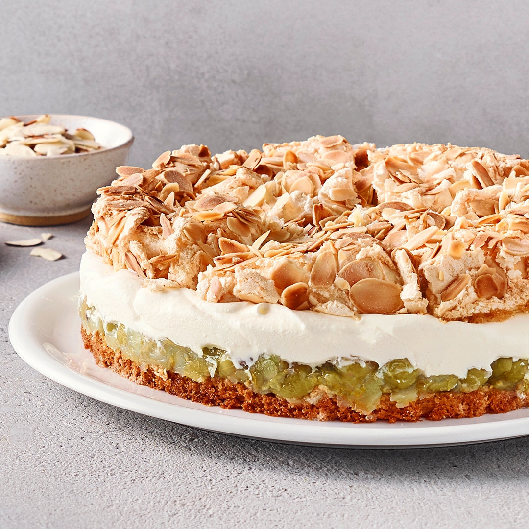 Ein Teller mit einem runden Kuchen, mit braunem Boden, grüner Fruchtschicht, weißer Cremeschicht und heller Knusperdecke.