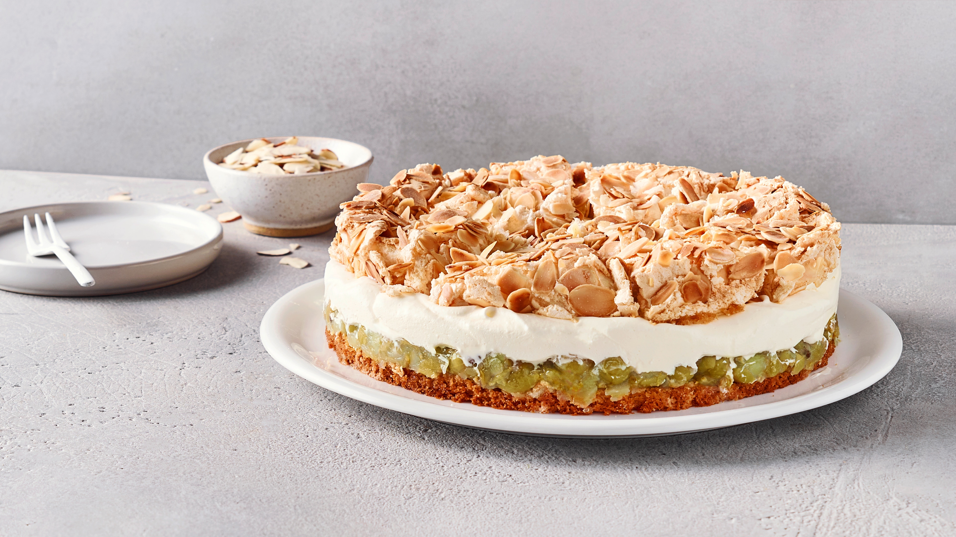 Ein Teller mit einem runden Kuchen, mit braunem Boden, grüner Fruchtschicht, weißer Cremeschicht und heller Knusperdecke.