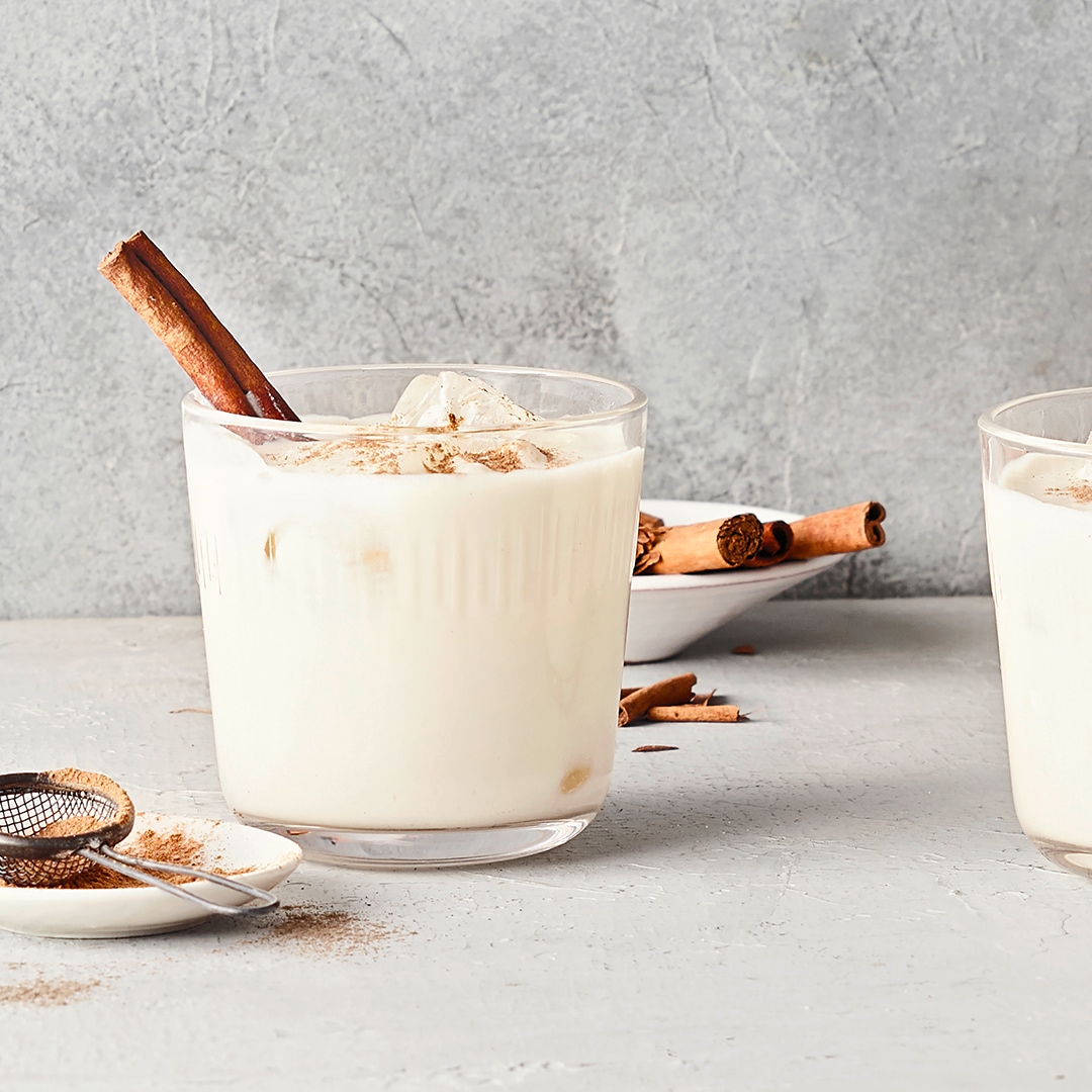 Ein Glas mit hellem Getränk, bestreut mit braunem Pulver und garniert mit einer Zimtstange, daneben ein Sieb mit Pulver