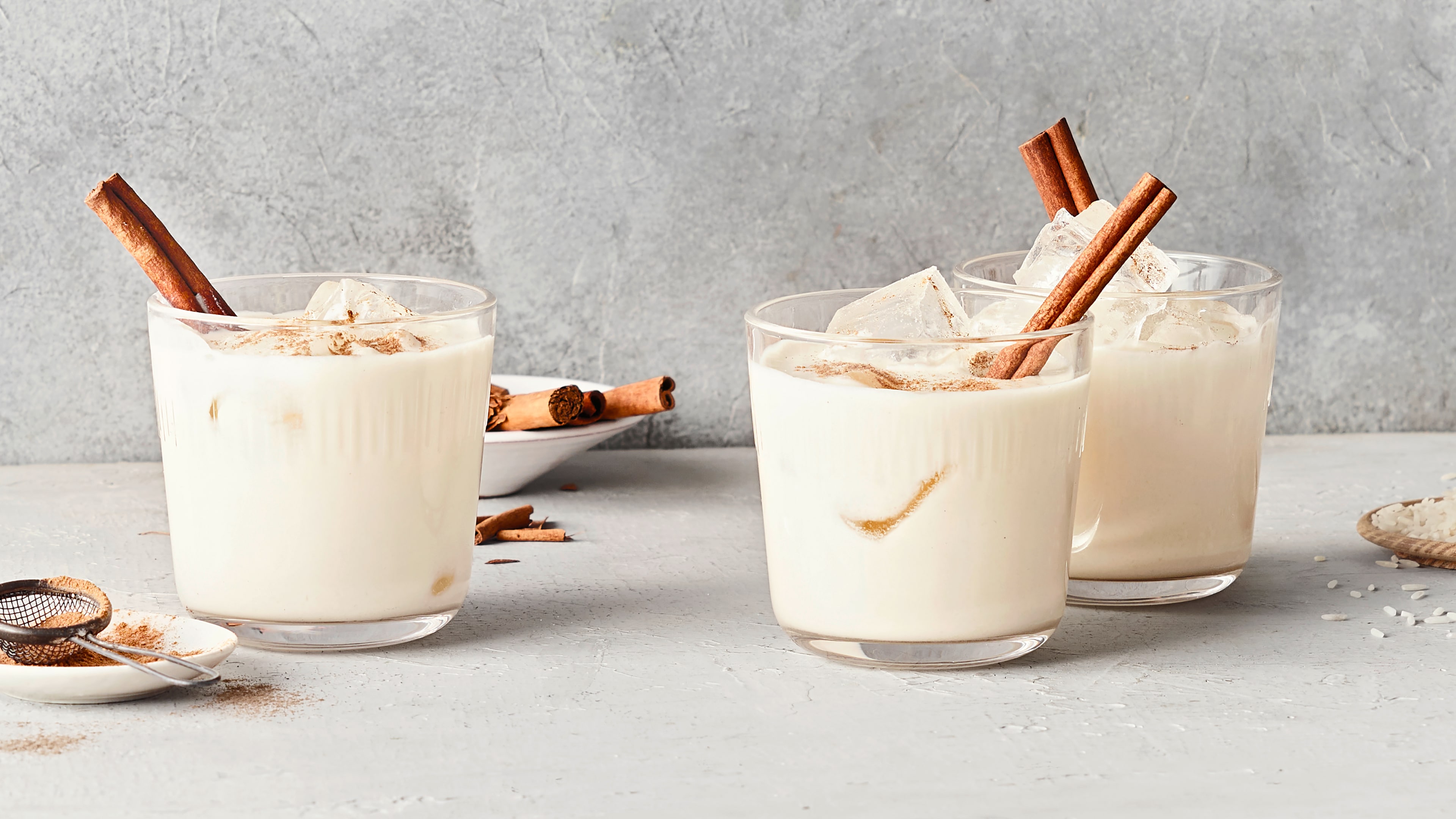 Ein Glas mit hellem Getränk, bestreut mit braunem Pulver und garniert mit einer Zimtstange, daneben ein Sieb mit Pulver