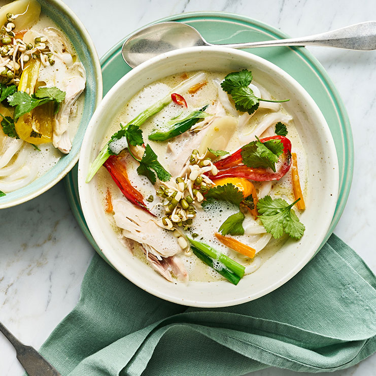 Eine Schüssel mit heller Suppe, darin Hühnerstücke, rote und gelbe Paprika, grüne Kräuter und Sprossen.