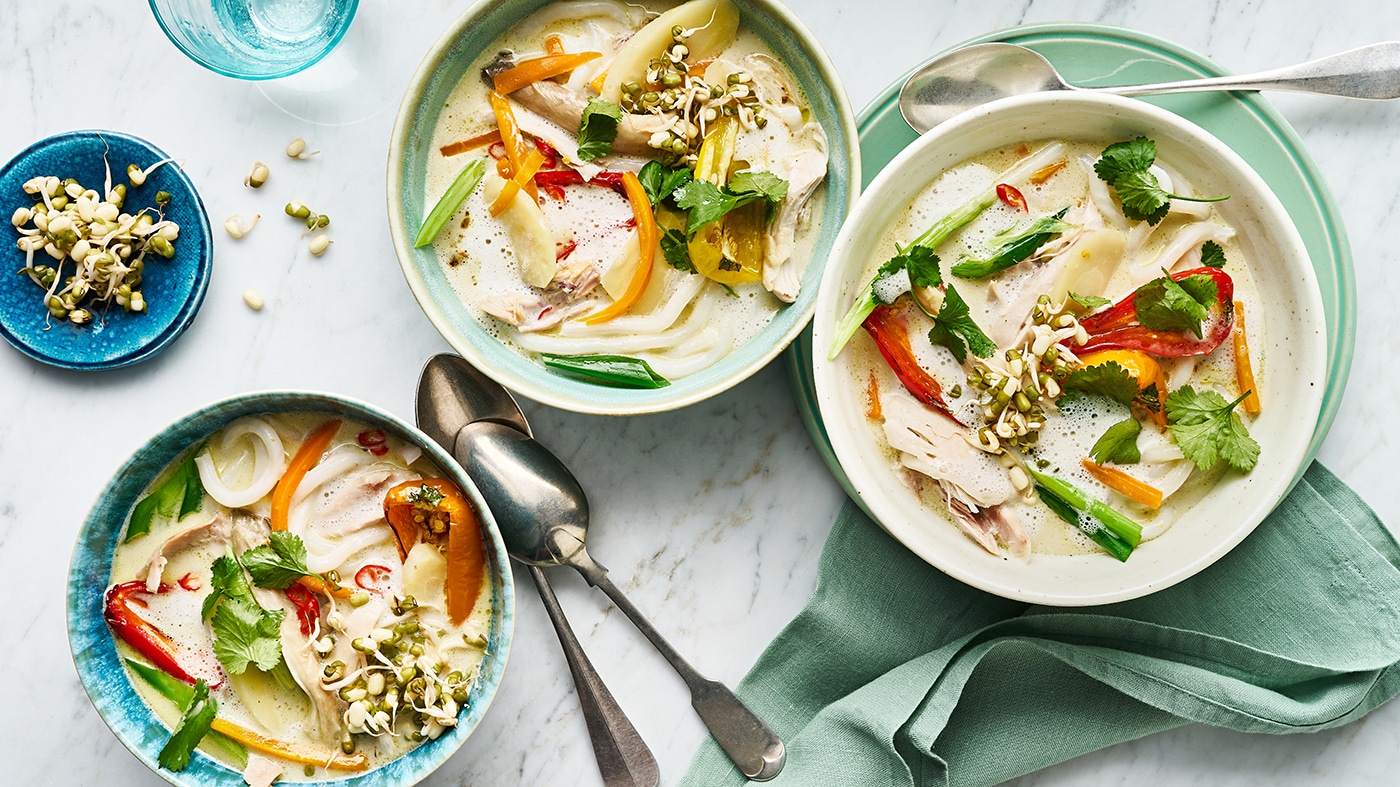 Eine Schüssel mit heller Suppe, darin Hühnerstücke, rote und gelbe Paprika, grüne Kräuter und Sprossen.