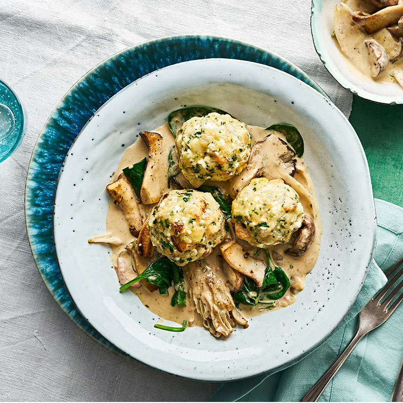 Ein Teller mit drei runden Knödeln in heller Soße, dazu braune Pilzstücke und grüne Spinatblätter.