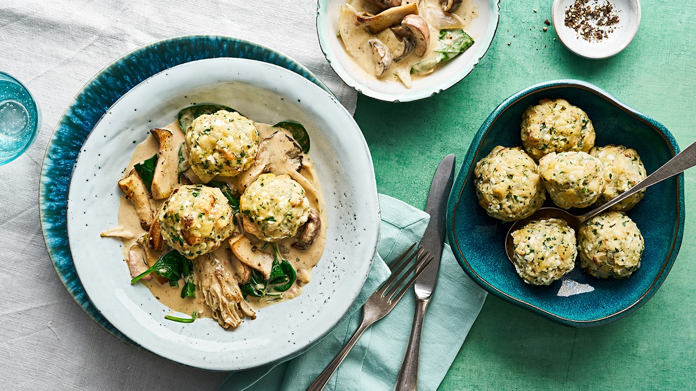 Ein Teller mit drei runden Knödeln in heller Soße, dazu braune Pilzstücke und grüne Spinatblätter.