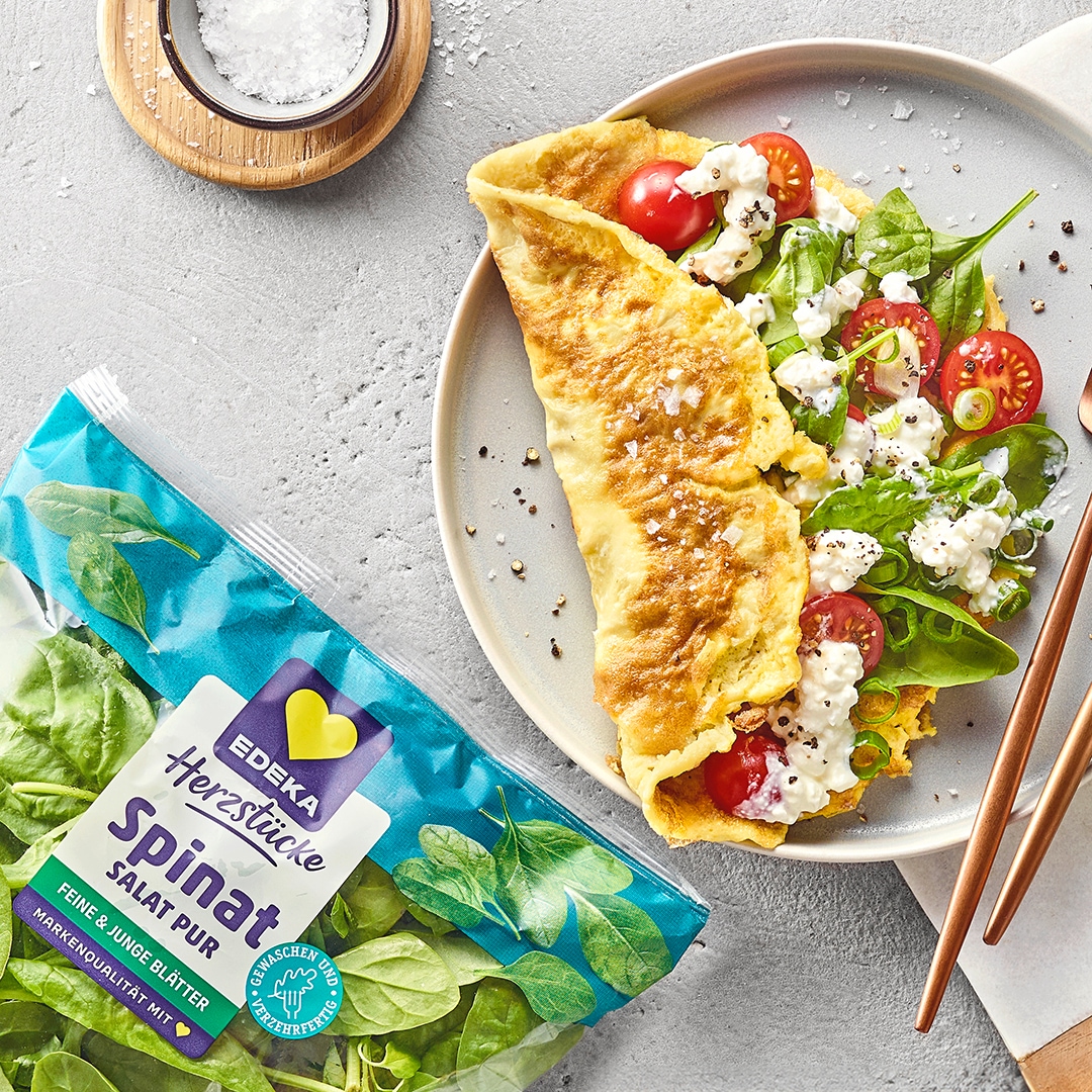 Omelett mit Tomaten und Hüttenkäse auf einem Teller, serviert mit Salat und Besteck. Daneben EDEKA Herzstücke Spinat Salat pur.