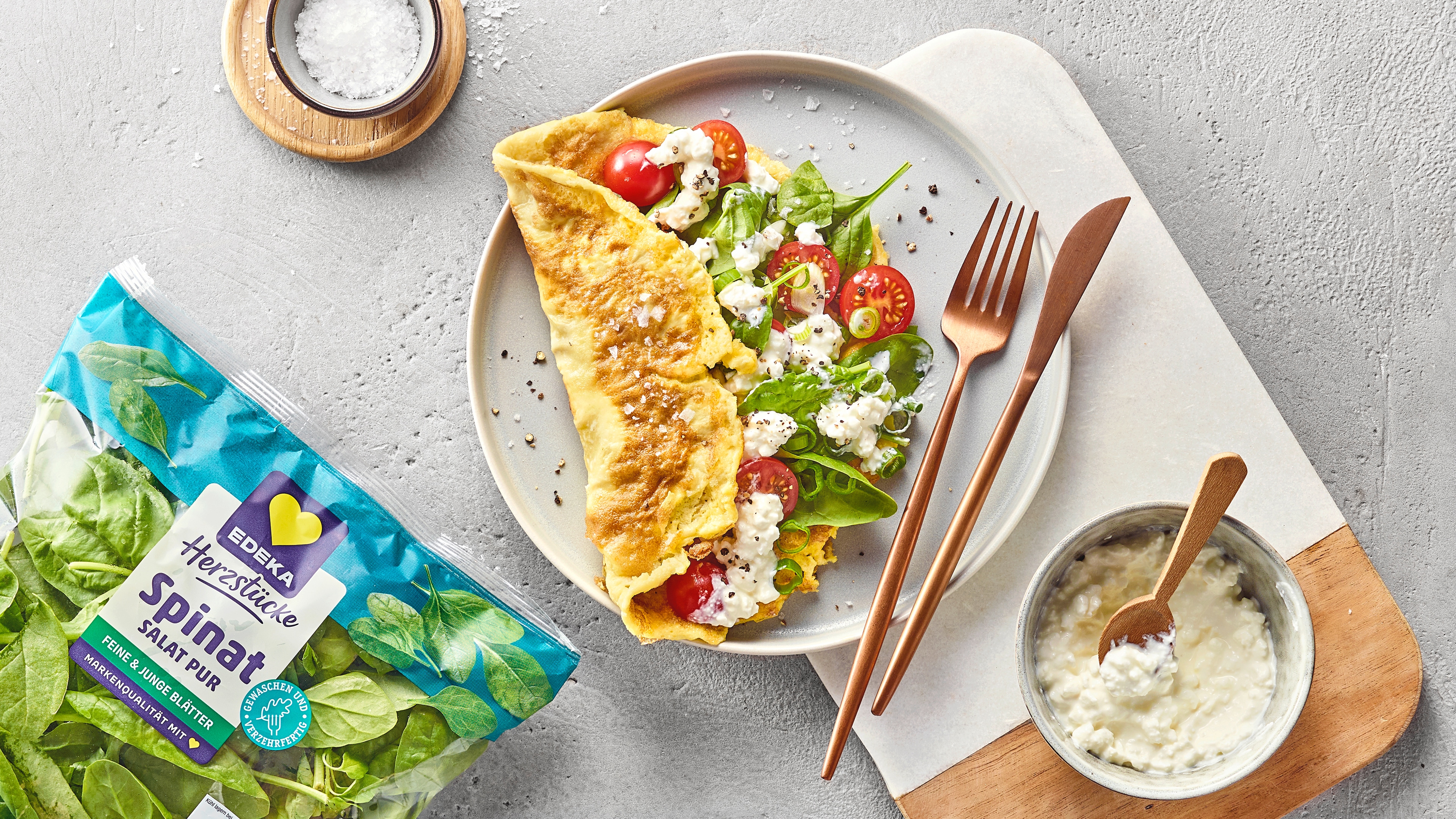 Omelett mit Tomaten und Hüttenkäse auf einem Teller, serviert mit Salat und Besteck. Daneben EDEKA Herzstücke Spinat Salat pur.