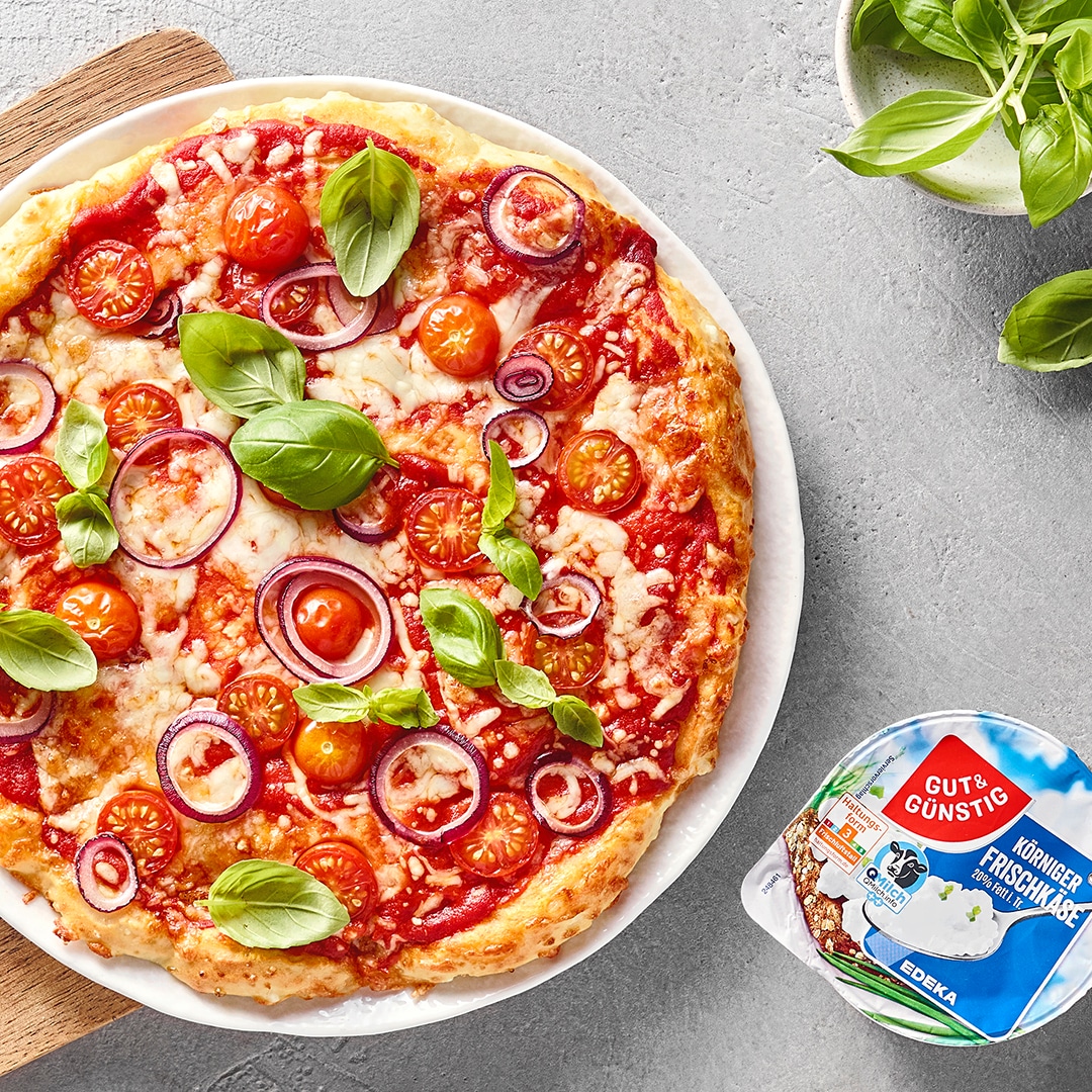 Pizza mit Tomatensauce, Käse, Kirschtomaten, roten Zwiebeln und frischem Basilikum auf einem Teller, daneben GUT&GÜNSTIG Körniger Frischkäse und eine Schale mit Basilikum.