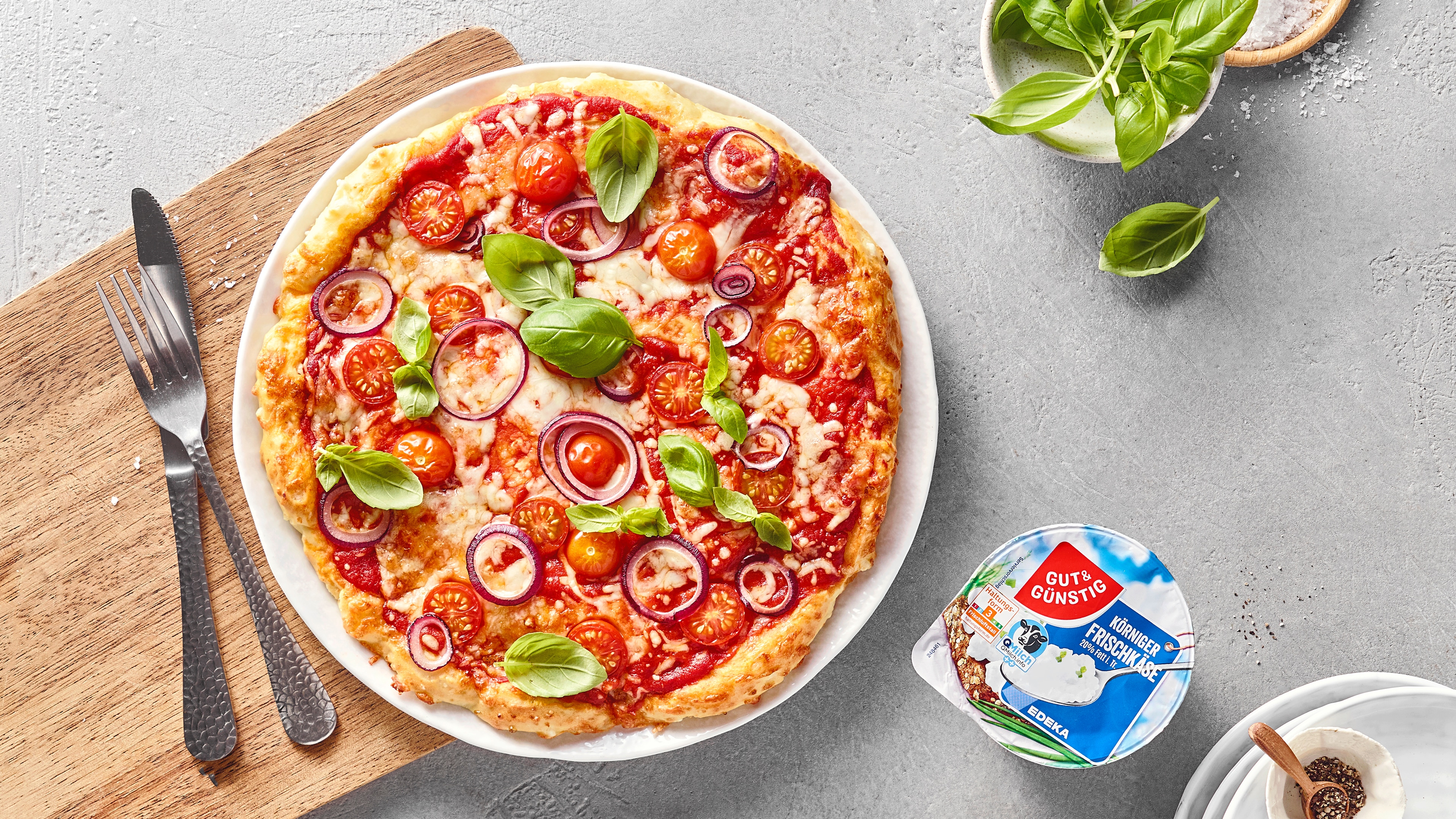 Pizza mit Tomatensauce, Käse, Kirschtomaten, roten Zwiebeln und frischem Basilikum auf einem Teller, daneben GUT&GÜNSTIG Körniger Frischkäse und eine Schale mit Basilikum.