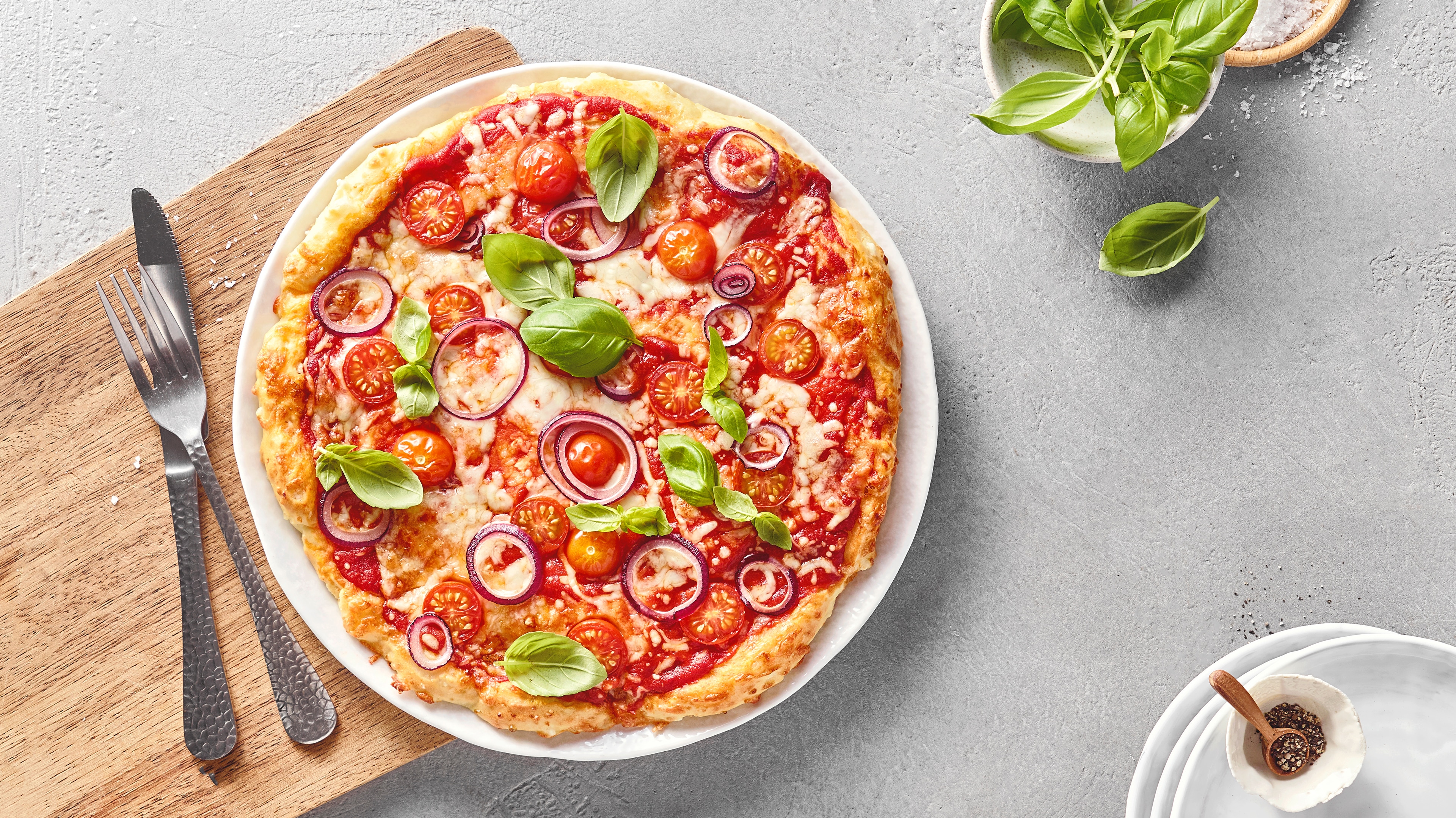 Pizza mit Tomatensauce, Käse, Kirschtomaten, roten Zwiebeln und frischem Basilikum auf einem Teller, daneben Besteck und eine Schale mit Basilikum.