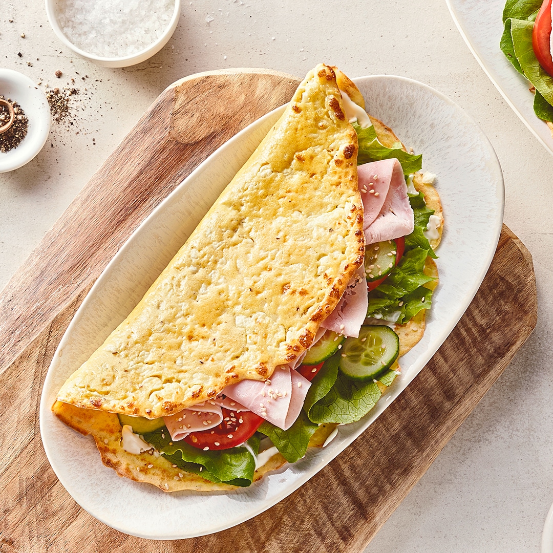 Ein Sandwich aus hellem, knusprigem Teig, gefüllt mit Salat, Fleischwust, Gurken und Tomatenscheiben, auf einem ovalen Teller