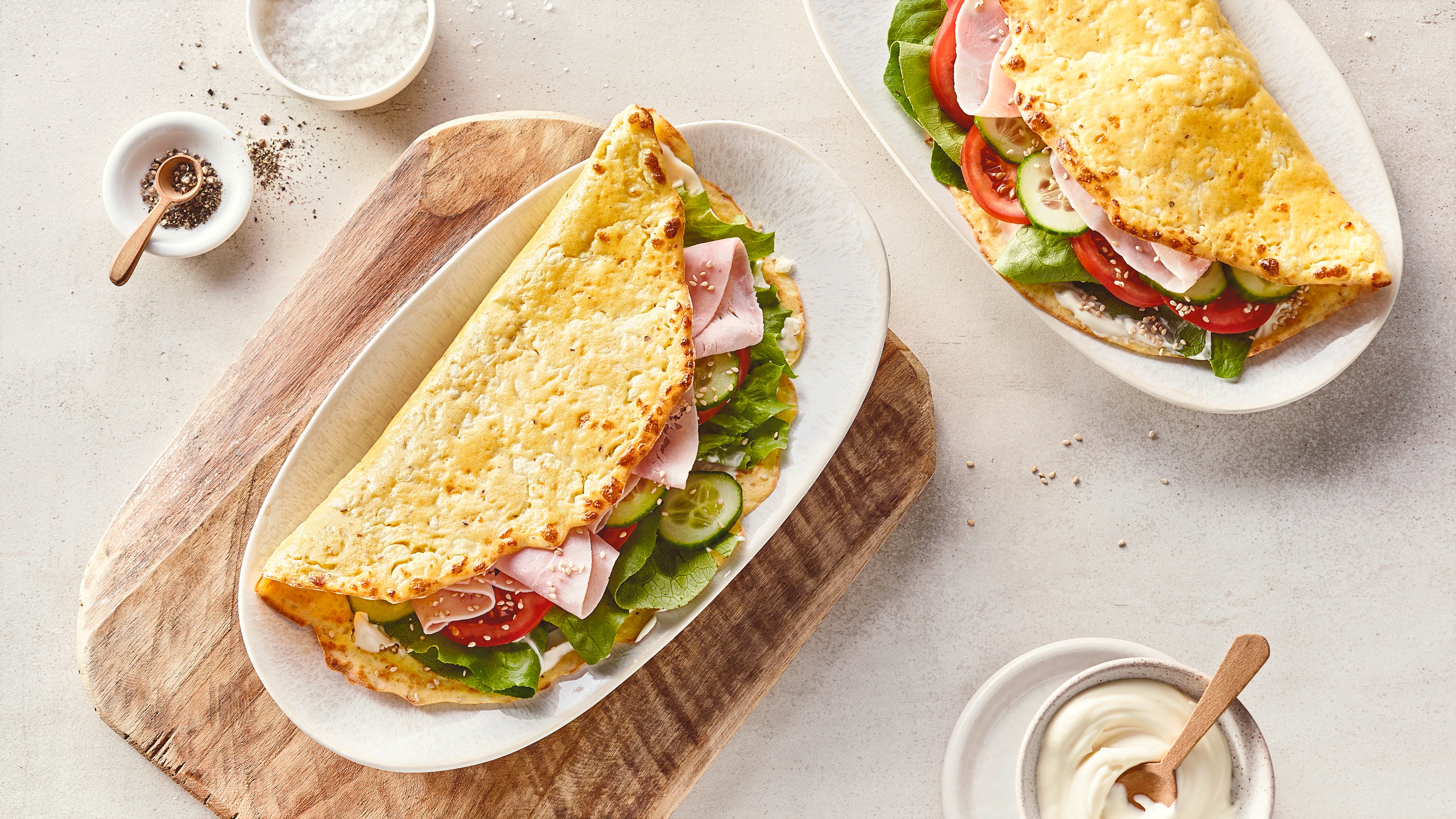Rezept für Hüttenkäse Sandwich Ein Sandwich aus hellem, knusprigem Teig, gefüllt mit Salat, Fleischwust, Gurken und Tomatenscheiben, auf einem ovalen Teller