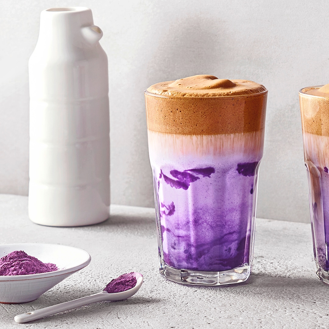Zwei Gläser mit lila Milchgetränk und cremigem Kaffeeschaum auf einer hellen Oberfläche, daneben eine weiße Flasche und lila Pulver.
