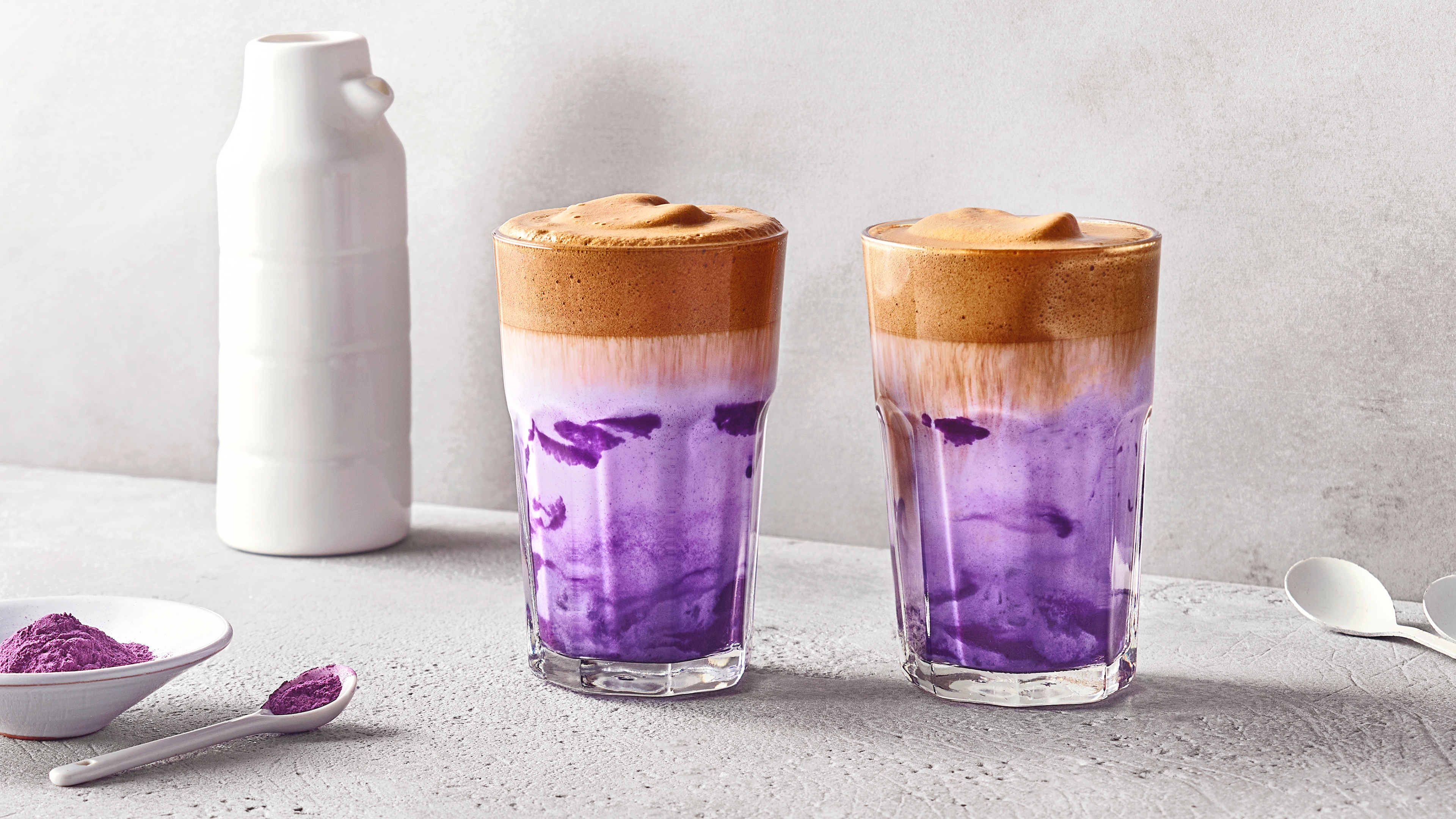 Zwei Gläser mit lila Milchgetränk und cremigem Kaffeeschaum auf einer hellen Oberfläche, daneben eine weiße Flasche und lila Pulver.
