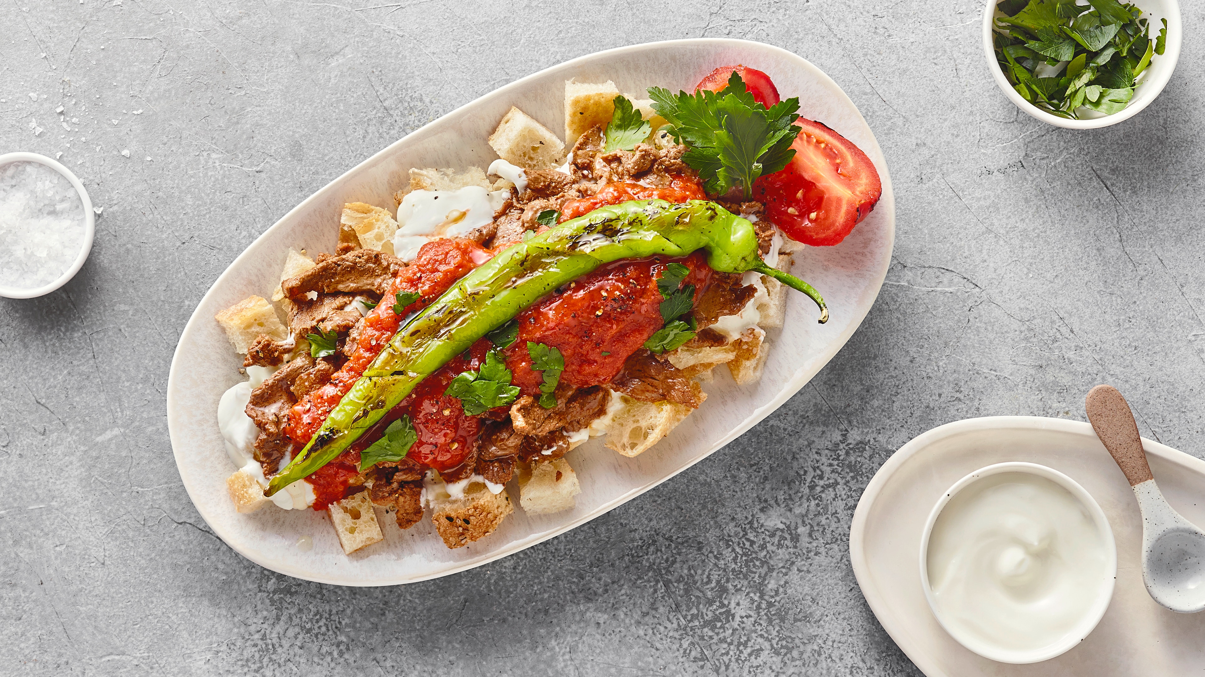 Rezept für Iskender Kebap Ein Teller mit Fleischstücken, Brotwürfeln und roter Soße, darauf eine grüne Pfefferschote, dazu Tomaten & Petersilie