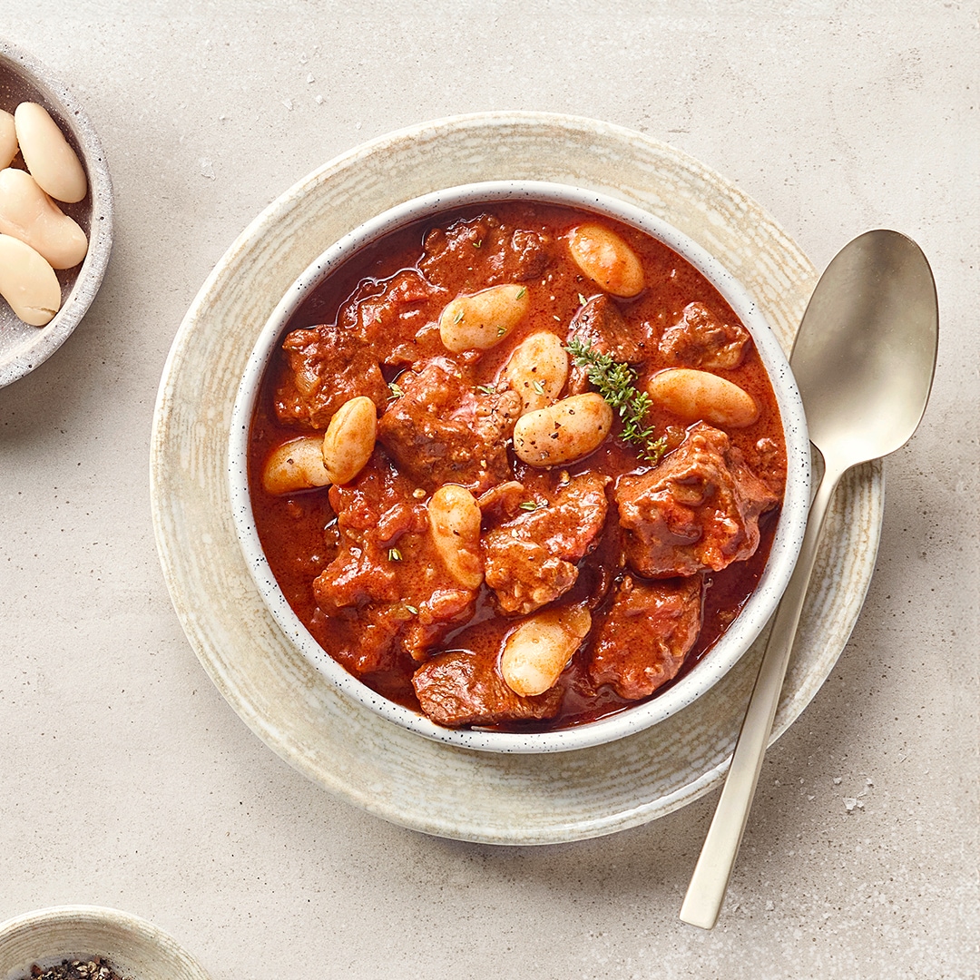 Eine Schüssel mit rotem Eintopf aus braunen Fleischstücken, weißen Bohnen und frischen Kräutern.