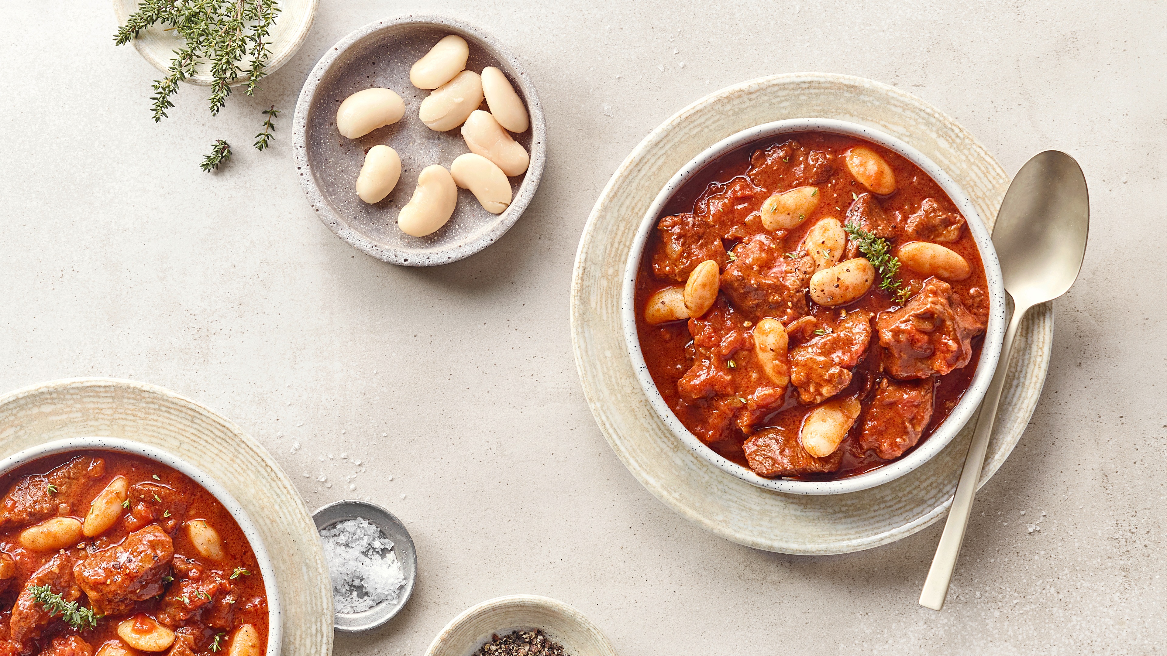 Eine Schüssel mit rotem Eintopf aus braunen Fleischstücken, weißen Bohnen und frischen Kräutern.