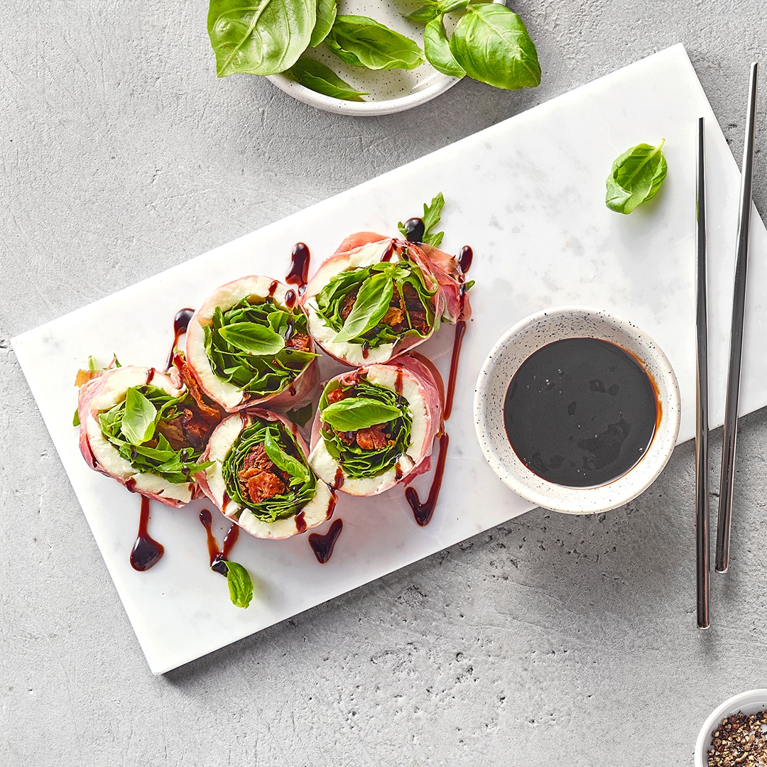 Sushi‑ähnliche Röllchen mit frischem Basilikum und dunkler Sauce auf Tellern, daneben Dip und Essstäbchen.