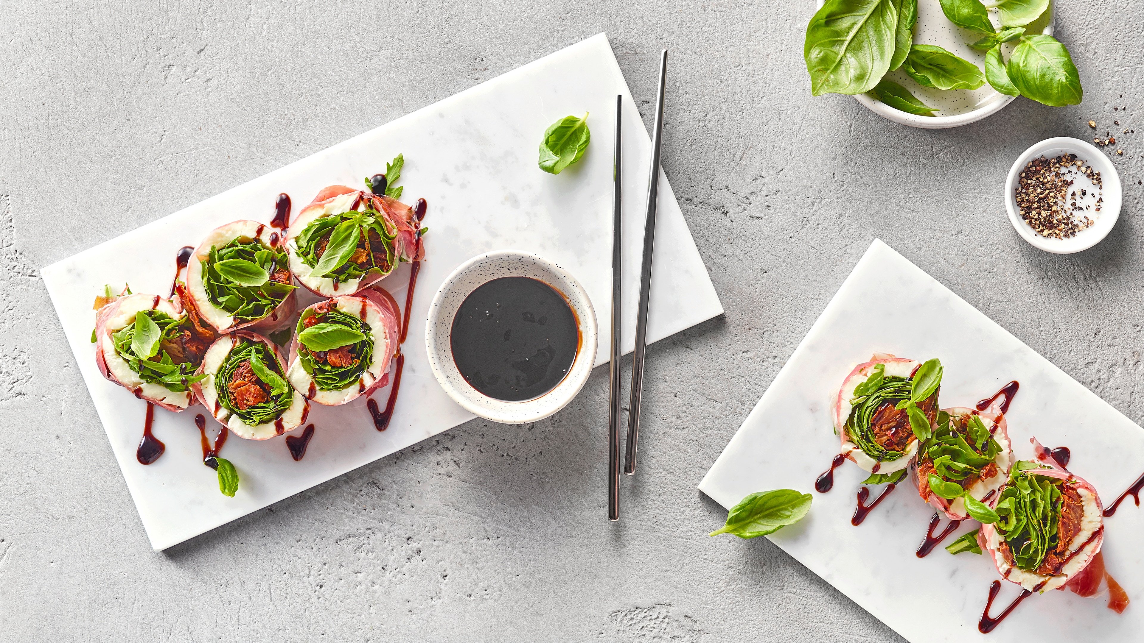 Sushi‑ähnliche Röllchen mit frischem Basilikum und dunkler Sauce auf Tellern, daneben Dip und Essstäbchen.