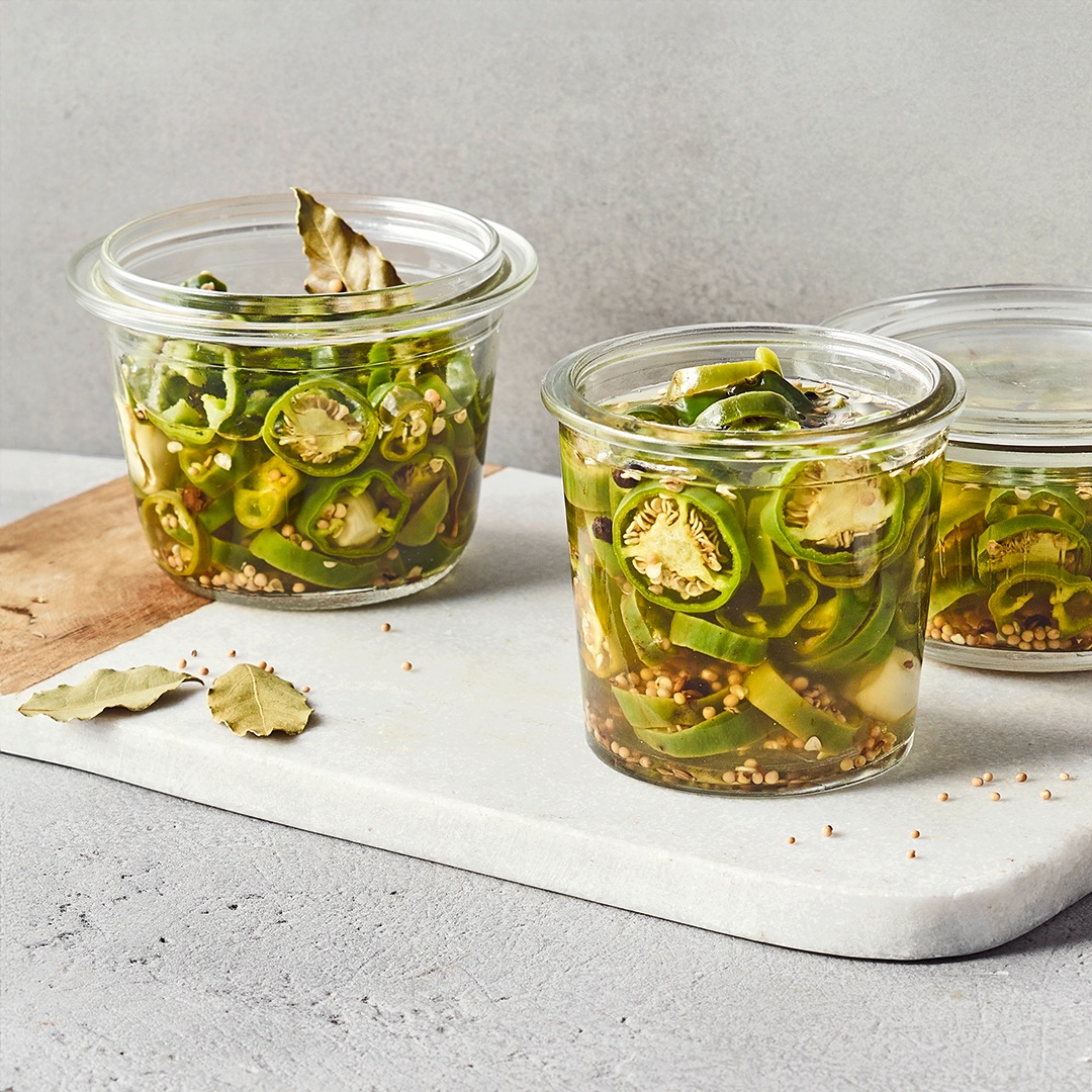 Ein Glas mit eingelegten grünen Chilischeiben in klarer Flüssigkeit, darin Gewürzkörner und Lorbeerblätter.