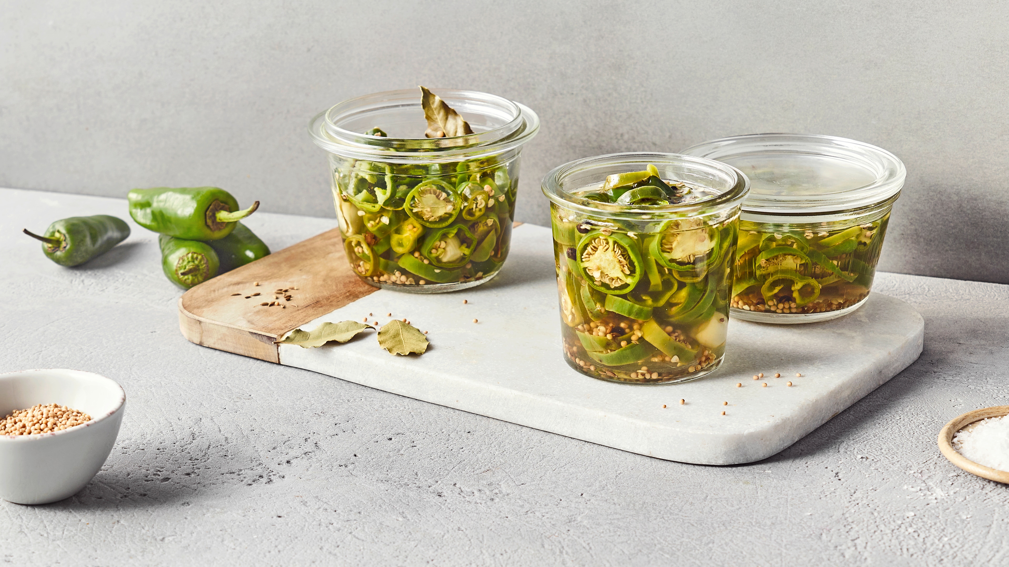 Ein Glas mit eingelegten grünen Chilischeiben in klarer Flüssigkeit, darin Gewürzkörner und Lorbeerblätter.