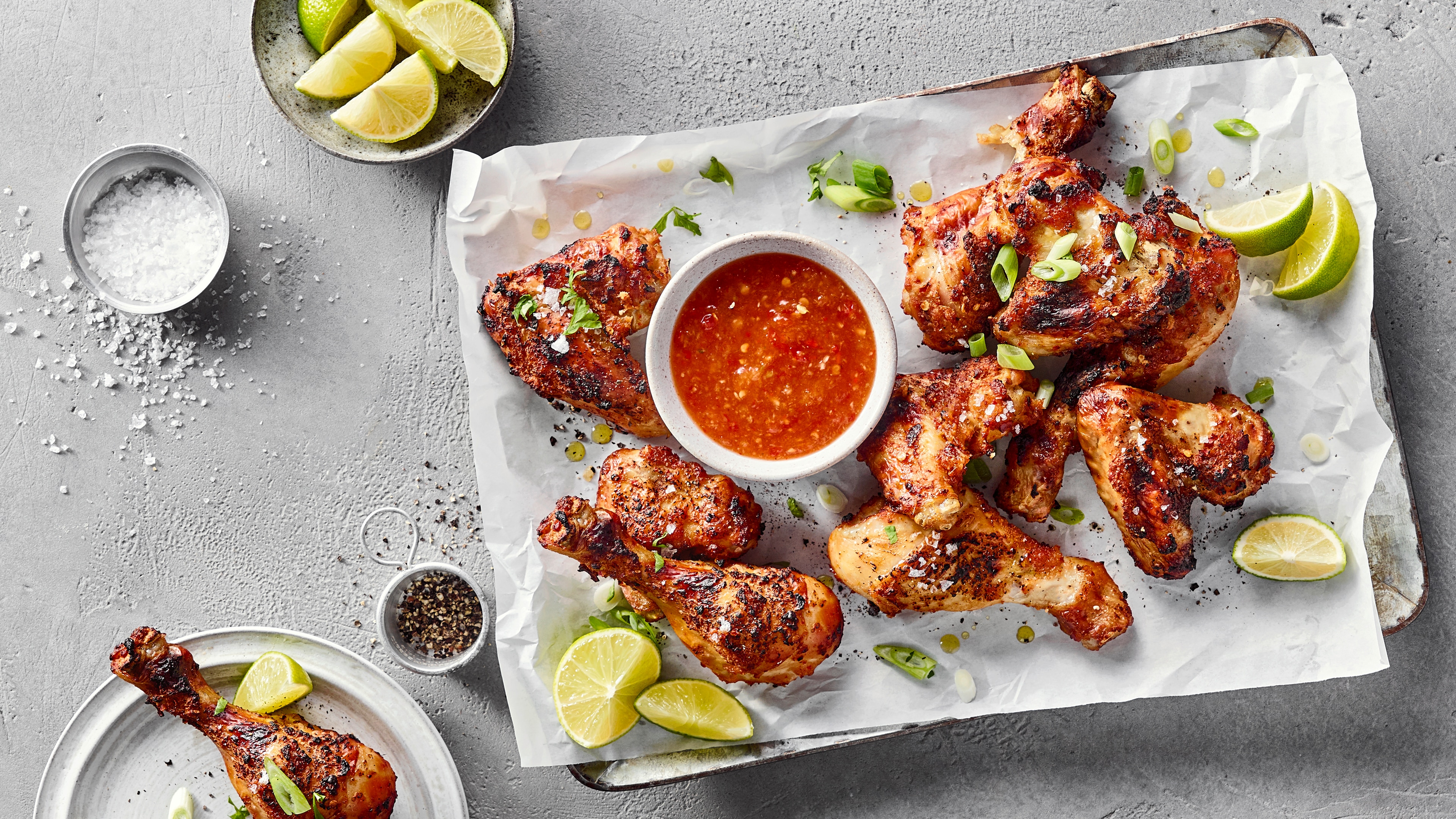 Ein Blech mit gebratenen Hähnchenteilen, grünen Zwiebelstücken, Limettenspalten und einer Schale mit roter Soße in der Mitte.