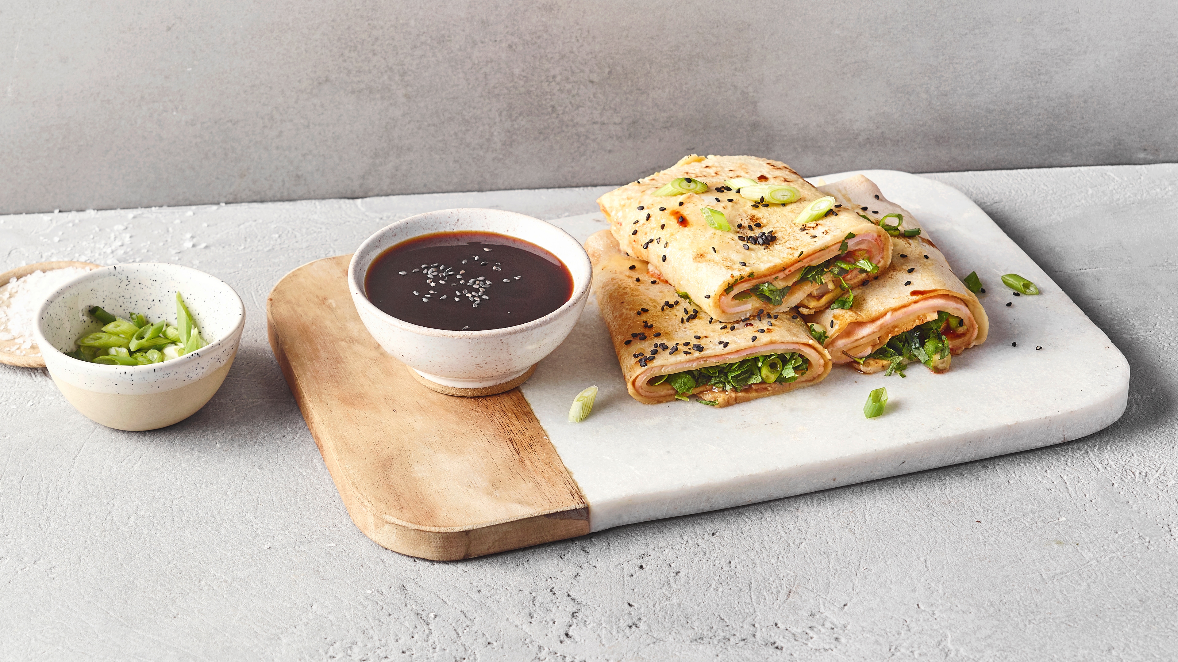 Ein Brett mit gefüllten Pfannkuchenrollen, darin frische Kräuter & Frühlingszwiebeln, bestreut mit Sesam, daneben eine Schale Soße.