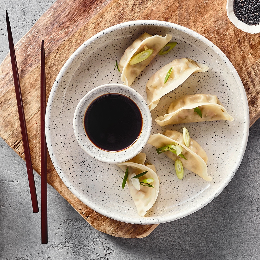 Eine Schüssel mit fünf hellen Teigtaschen, garniert mit grünen Zwiebelringen, und einer Schale dunkler Soße in der Mitte.
