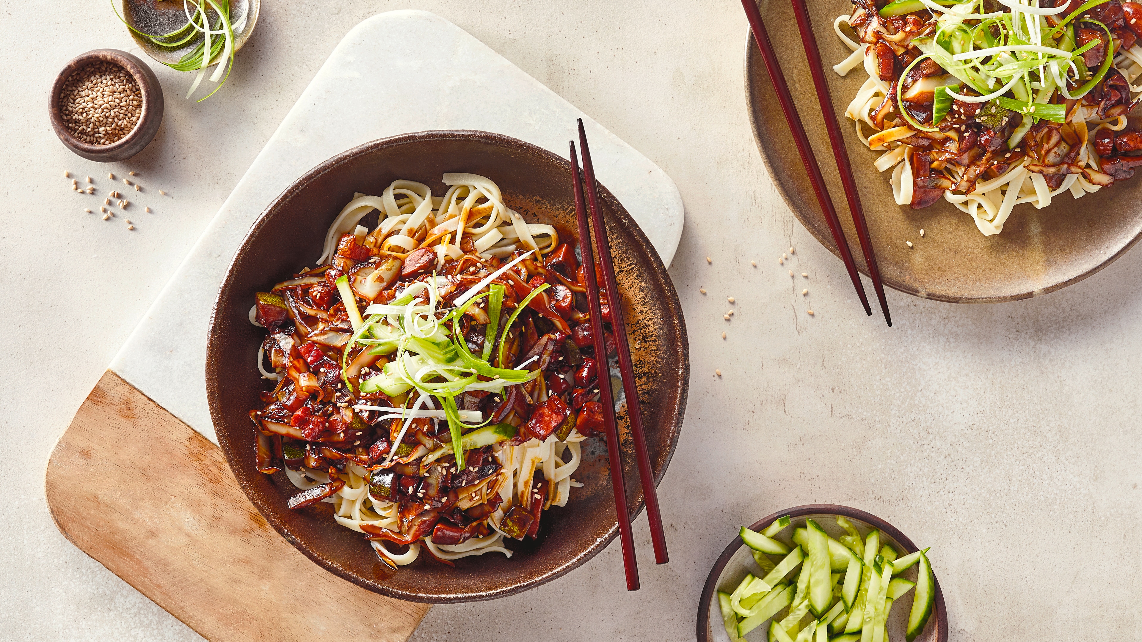 Koreanisches Nudel-Rezept für Jjajangmyeon Eine Schale mit breiten Reisnudeln und Zucchini, in bräunlicher Soße, getoppt mit Gurkenstreifen, Gurke und Sesam