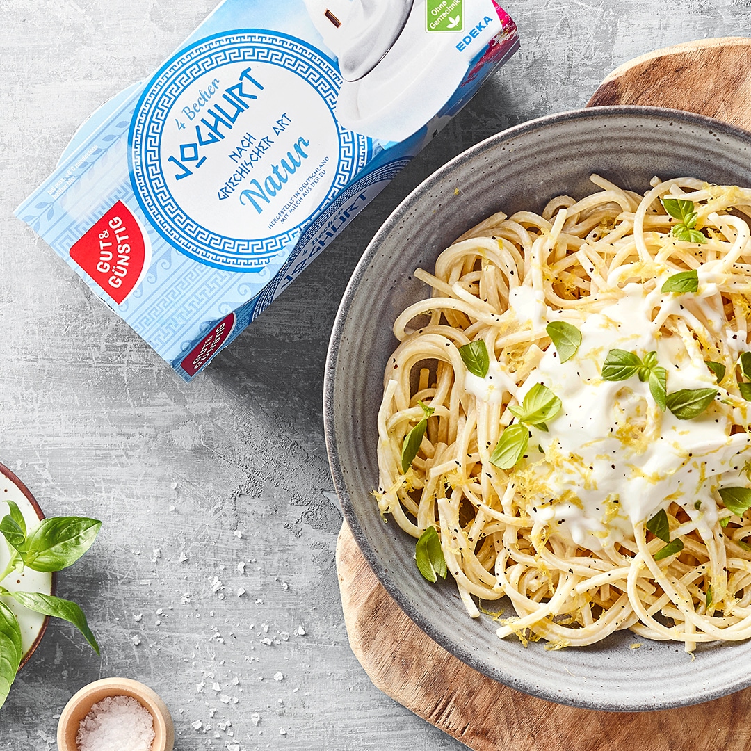 Spaghetti mit cremiger Joghurtsauce und frischem Basilikum in einer Schüssel, angerichtet auf einem Holzbrett, daneben GUT&GÜNSTIG griechischer Joghurt.
