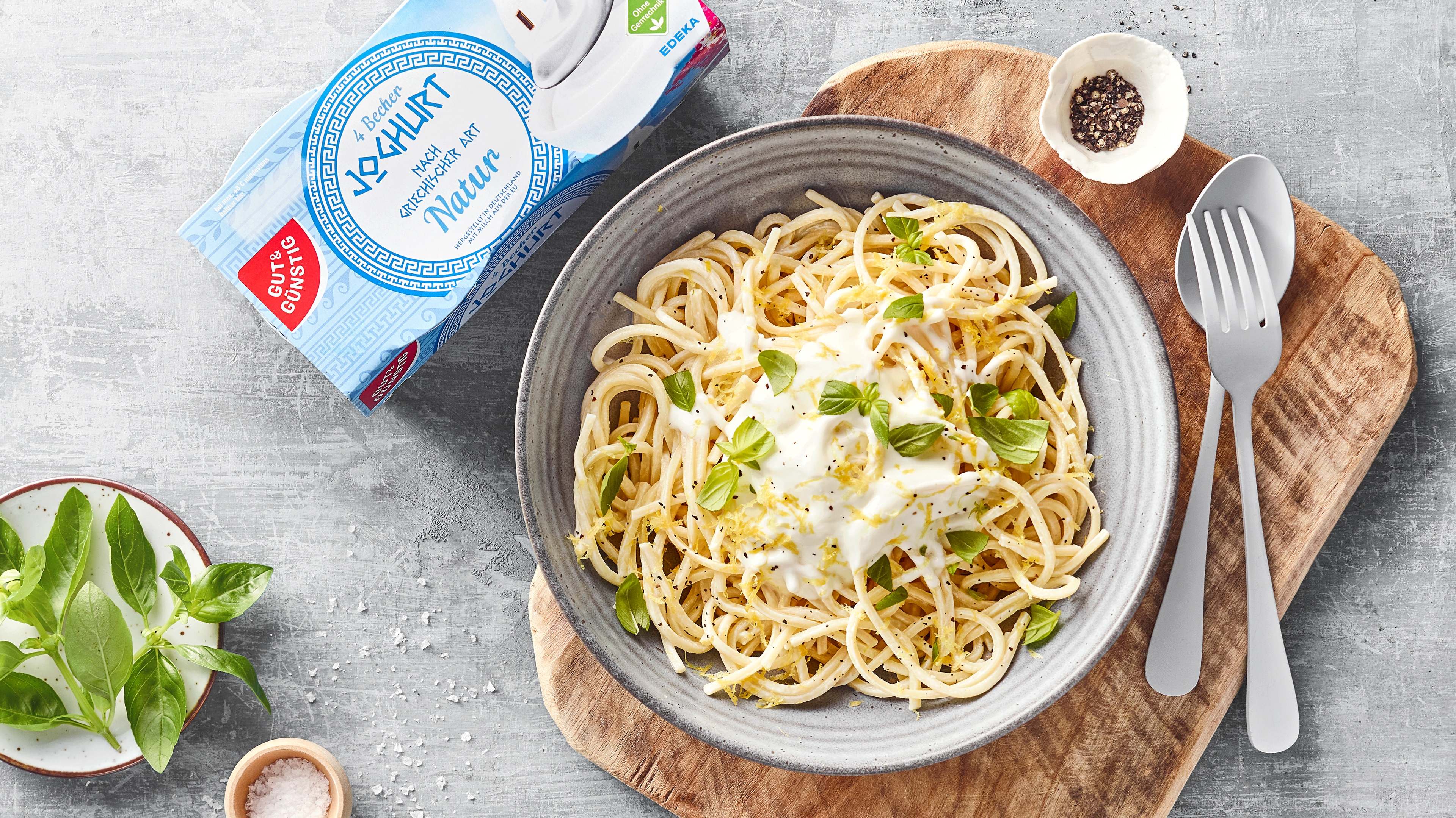 Spaghetti mit cremiger Joghurtsauce und frischem Basilikum in einer Schüssel, angerichtet auf einem Holzbrett, daneben GUT&GÜNSTIG griechischer Joghurt.