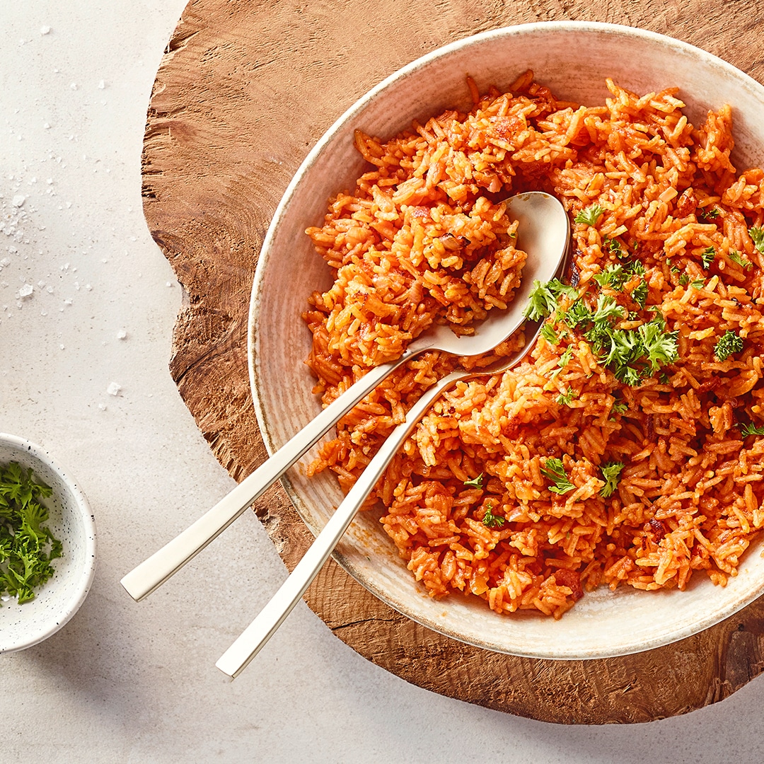 Eine Schüssel mit Reis in roter Soße, darauf grüne Kräuter, daneben zwei silberne Löffel auf Holzunterlage.