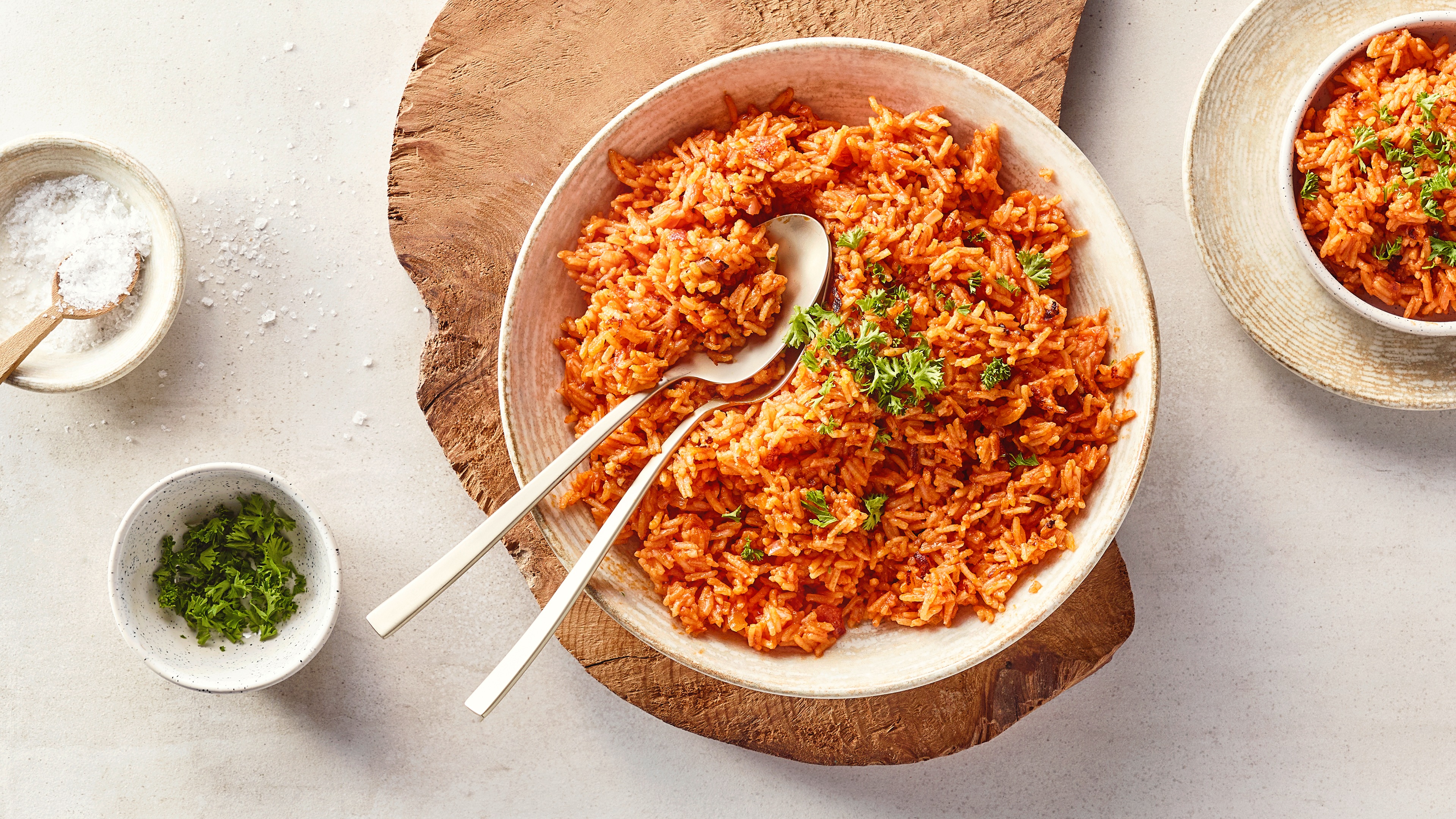 Eine Schüssel mit Reis in roter Soße, darauf grüne Kräuter, daneben zwei silberne Löffel auf Holzunterlage.