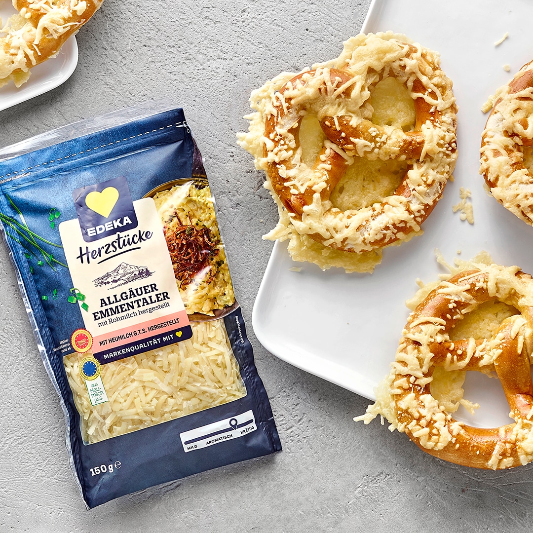 Goldbraun gebackene Laugenbrezeln, mit geschmolzenem Käse überzogen. Links daneben EDEKA Herzstücke Allgäuer Emmentaler.