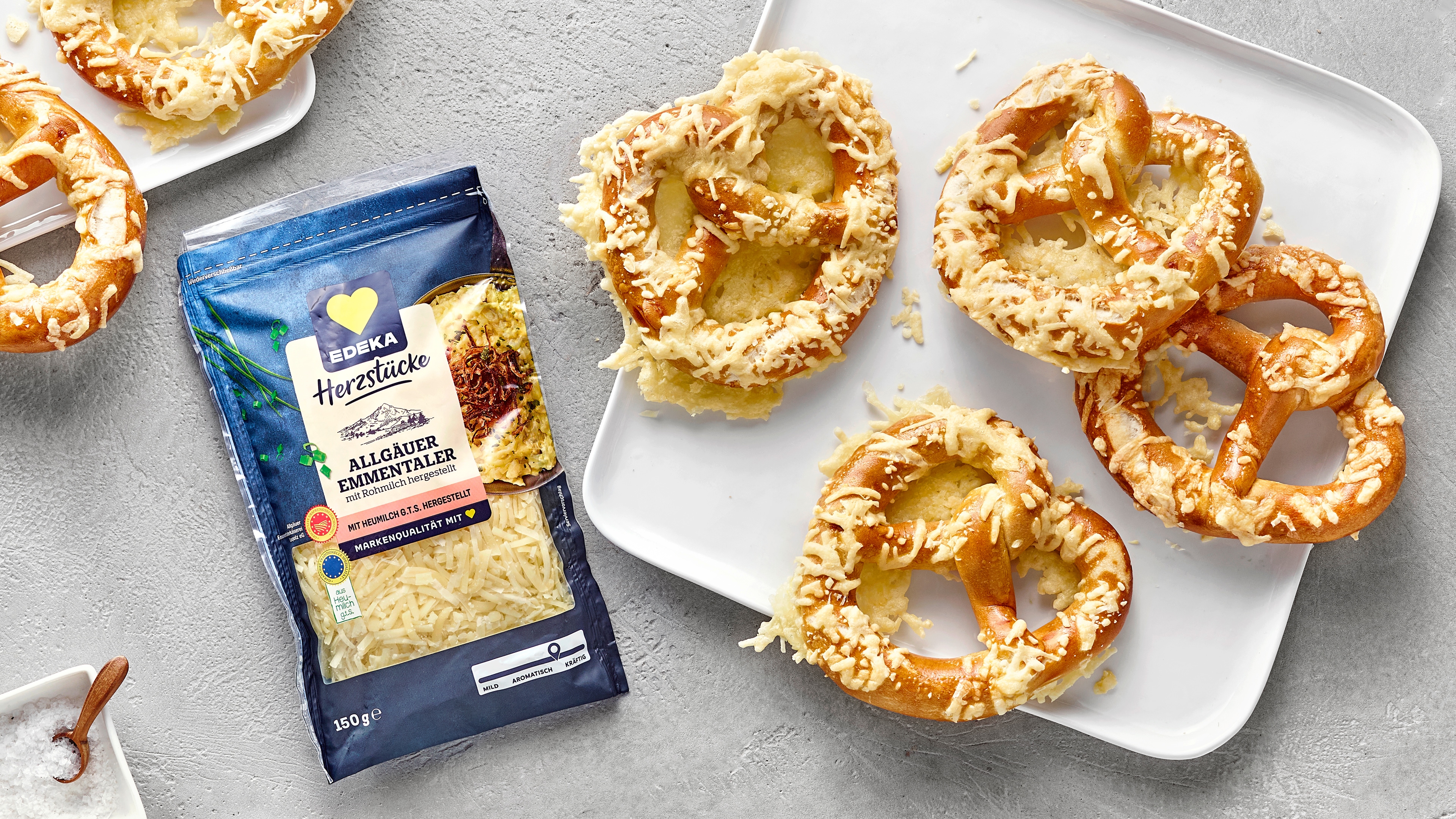 Goldbraun gebackene Laugenbrezeln, mit geschmolzenem Käse überzogen. Links daneben EDEKA Herzstücke Allgäuer Emmentaler.